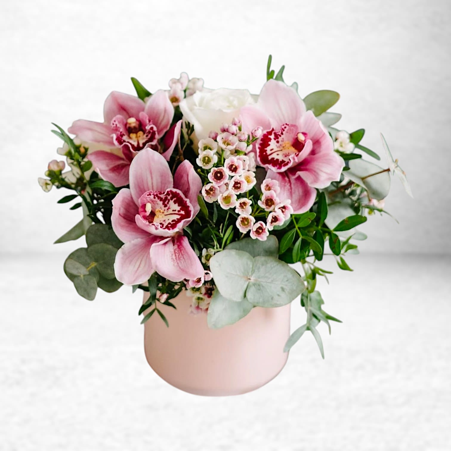 Orchid Blush Bloom - Elevate any moment with the delicate beauty of Orchid Blush Bloom. This compact arrangement features soft pink cymbidium orchids, petite blush waxflower, creamy white roses, and elegant sprigs of eucalyptus—artfully styled in a modern matte blush ceramic vase.  Perfect for birthdays, thank-you gifts, or just because, this petite yet luxurious floral design is available for same-day delivery in Galveston, South Houston, League City, and surrounding areas. It’s a lovely gesture of refined charm and thoughtfulness.  Substitution Policy: Cymbidium orchids are seasonal and may be substituted with similar premium blooms like ranunculus or garden roses if unavailable. White roses may be replaced with comparable ivory or soft blush alternatives. Vase style may vary slightly depending on availability. All substitutions will maintain the original style, value, and color palette.