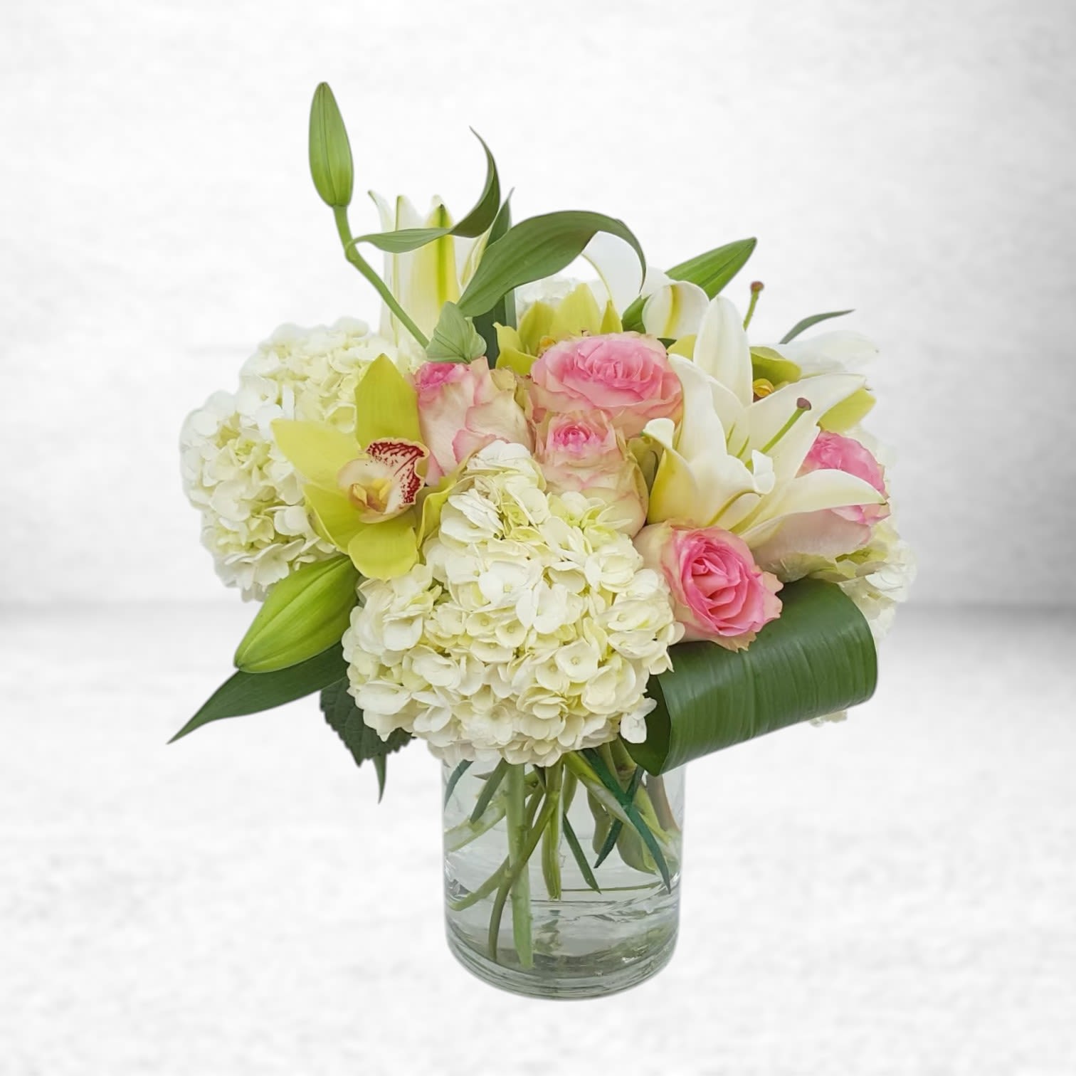Lily Crème Reverence - Graceful and refined, Lily Crème Reverence is a luxury floral arrangement designed to impress. Featuring voluminous white hydrangea, soft pink garden roses, lush oriental lilies, and exotic green cymbidium orchids, this design radiates purity and elegance with a romantic twist. Accented with sculptural tropical foliage and arranged in a tall clear cylinder vase, it’s perfect for anniversaries, sympathy flower delivery in Houston, or as a statement piece for corporate gifting or weddings.  Same-day delivery available across Galveston, League City, Dickinson, Friendswood  Substitution Policy To ensure the freshest blooms possible, substitutions may be made depending on seasonal availability. If white lilies, garden roses, or orchids are unavailable, we will replace them with premium florals in similar shades and style to maintain the design's integrity and value.  