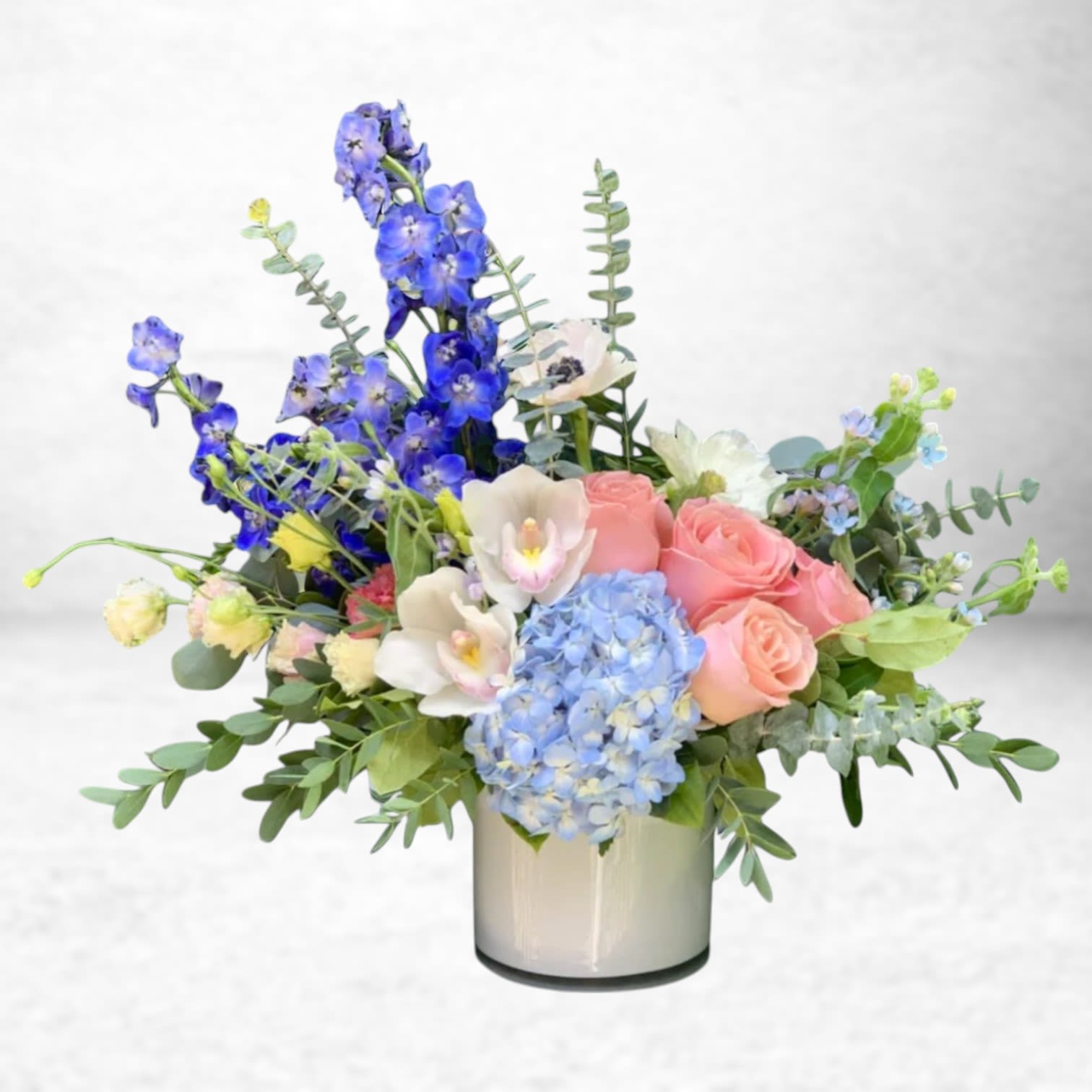 Skyline Reverie - Let your thoughts drift with Skyline Reverie, a dreamy arrangement featuring pastel pink roses, delicate white cymbidium orchids, airy blue hydrangea, and striking delphinium in shades of sapphire and indigo. Designed with a modern silhouette and overflowing with fresh eucalyptus and textural accents, this bouquet evokes the calm and wonder of a Galveston sunset.