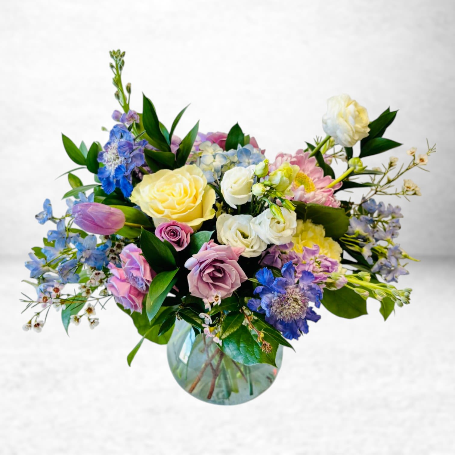 Meadowlight Breeze - Meadowlight Breeze evokes the feeling of a soft spring morning, filled with color, fragrance, and joy. This airy garden-style arrangement blends pastel roses, blue delphinium, lavender lisianthus, yellow garden roses, and tulips with lush greenery. Thoughtfully arranged in a clear glass vase, it brings a delicate yet uplifting burst of color to any setting.  Perfect for birthdays, new babies, get-well wishes, or simply celebrating life’s beautiful moments. This is a light-filled gesture that radiates calm and care.  Substitution Policy: We use only the freshest blooms available. In the rare event that certain flowers need to be substituted, we will maintain the arrangement’s overall color palette, composition, and value to reflect the spirit of the original design.