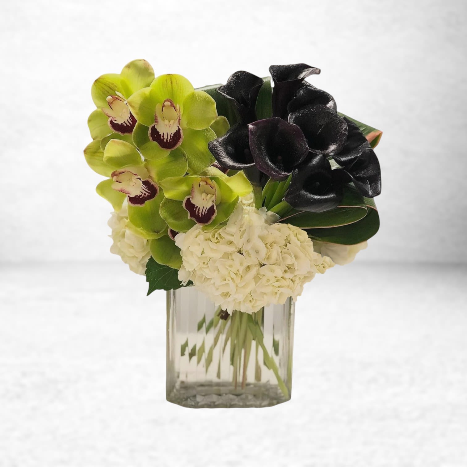 Nocturne Luxe - Bold and unexpected, Nocturne Luxe is a dramatic floral design that makes a powerful statement. Featuring velvety black calla lilies, vibrant green cymbidium orchids, and lush white hydrangea, this arrangement blends modern sophistication with natural elegance. Displayed in a tall, clear glass vase with faceted detailing, it’s ideal for upscale celebrations, corporate gifting, or anyone with a taste for the extraordinary.  This showstopping bouquet is available for delivery in Galveston, South Houston, and surrounding areas. Perfect for anniversaries, luxury events, or as a stunning sympathy piece with a modern edge.  Substitution Policy: Due to the rarity and seasonal nature of black calla lilies and cymbidium orchids, substitutions may be necessary. We will carefully select similar blooms—such as deep burgundy mini callas or green dendrobium orchids—to maintain the striking visual contrast and luxury aesthetic. Vase substitutions may apply but will match in height and style.
