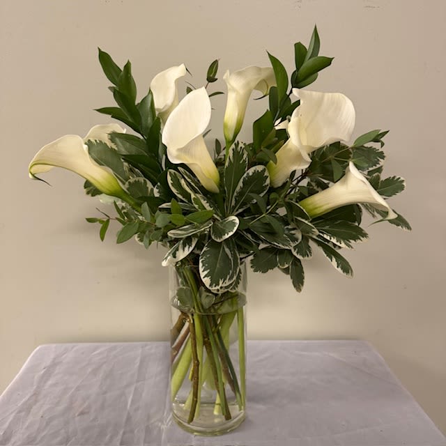 Charming Calla  - Pure white calla lilies arranged with sculptural grace-the Charming Calla is a white flower arrangement perfect for sympathy, a wedding, or any occasion that calls for calm elegance. The clean lines and serene silhouette make the recipient feel centered and seen; this is the kind of bouquet that says thoughtful, respectful, and beautifully simple.  Several long-stemmed white calla lilies are gathered upright in a clear cylindrical glass vase, accented by dense green foliage that provides rich contrast and a fresh, natural texture. The compact composition maintains a formal, restrained mood and fits well on a mantel, altar, or reception table. Vase included; stems are arranged for easy display and simple care-keep water fresh and trim stems as needed to preserve the lilies' sculptural beauty.  Please note:  Substitutions may be made to ensure the freshest product in your arrangement.