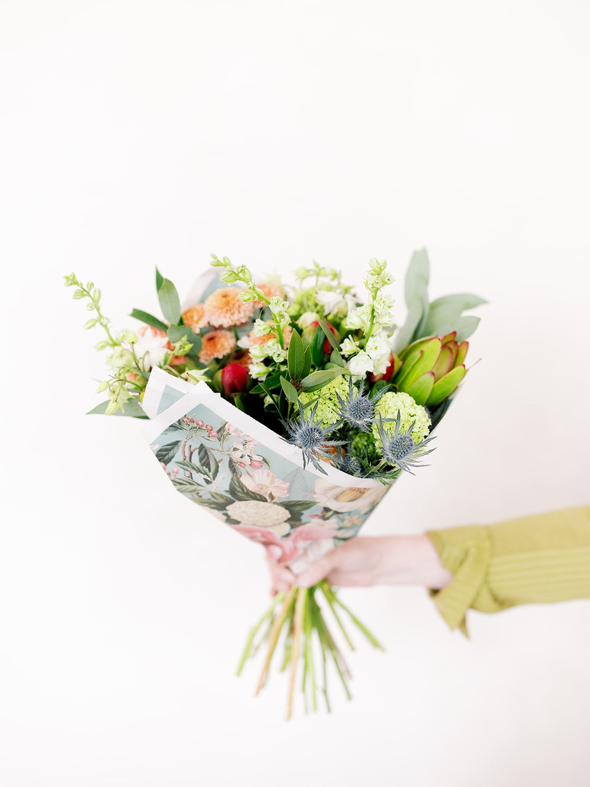 Handheld bouquet with peach, green, and blue flowers in floral wrap