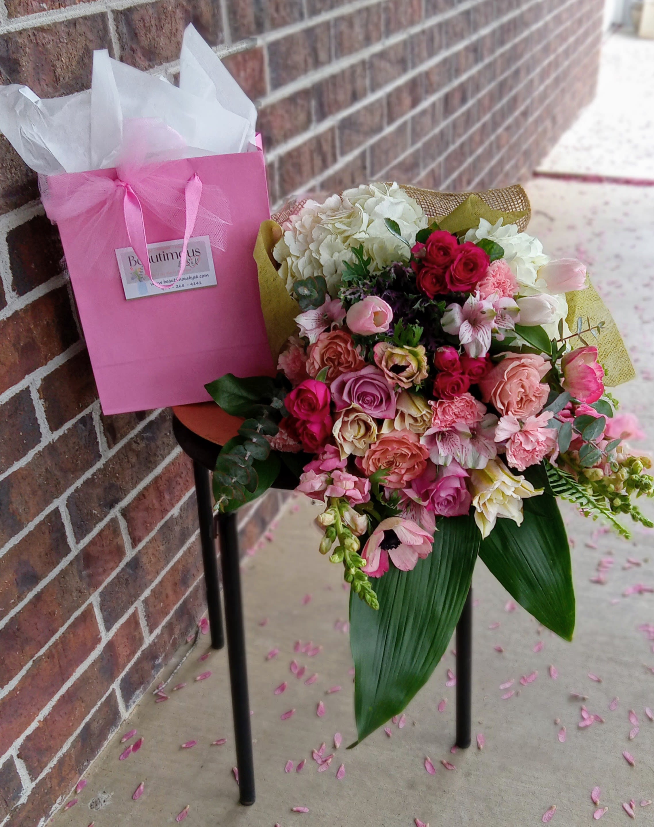 Pink and white mixed flower bouquet in a gift bag beside a brick wall