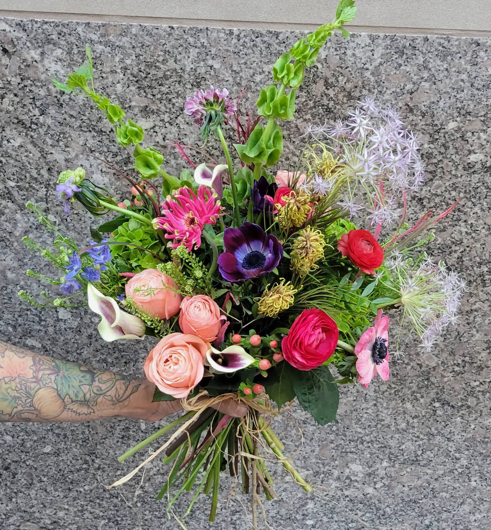 Wild Spring Meadow - This exquisite bouquet features elegant anemones, their delicate petals and dark centers adding a touch of sophistication. Complementing them are lush ranunculus blooms, known for their layered, rose-like appearance and rich, vibrant colors. Graceful calla lilies add an element of timeless elegance, their sleek, trumpet-shaped blooms standing out amidst the mix.