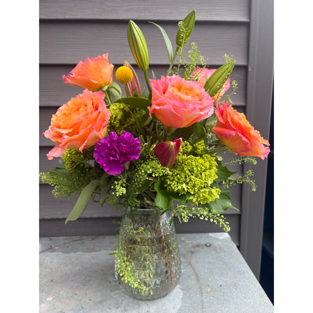 Orange and pink roses with lilies and green accents in a glass vase