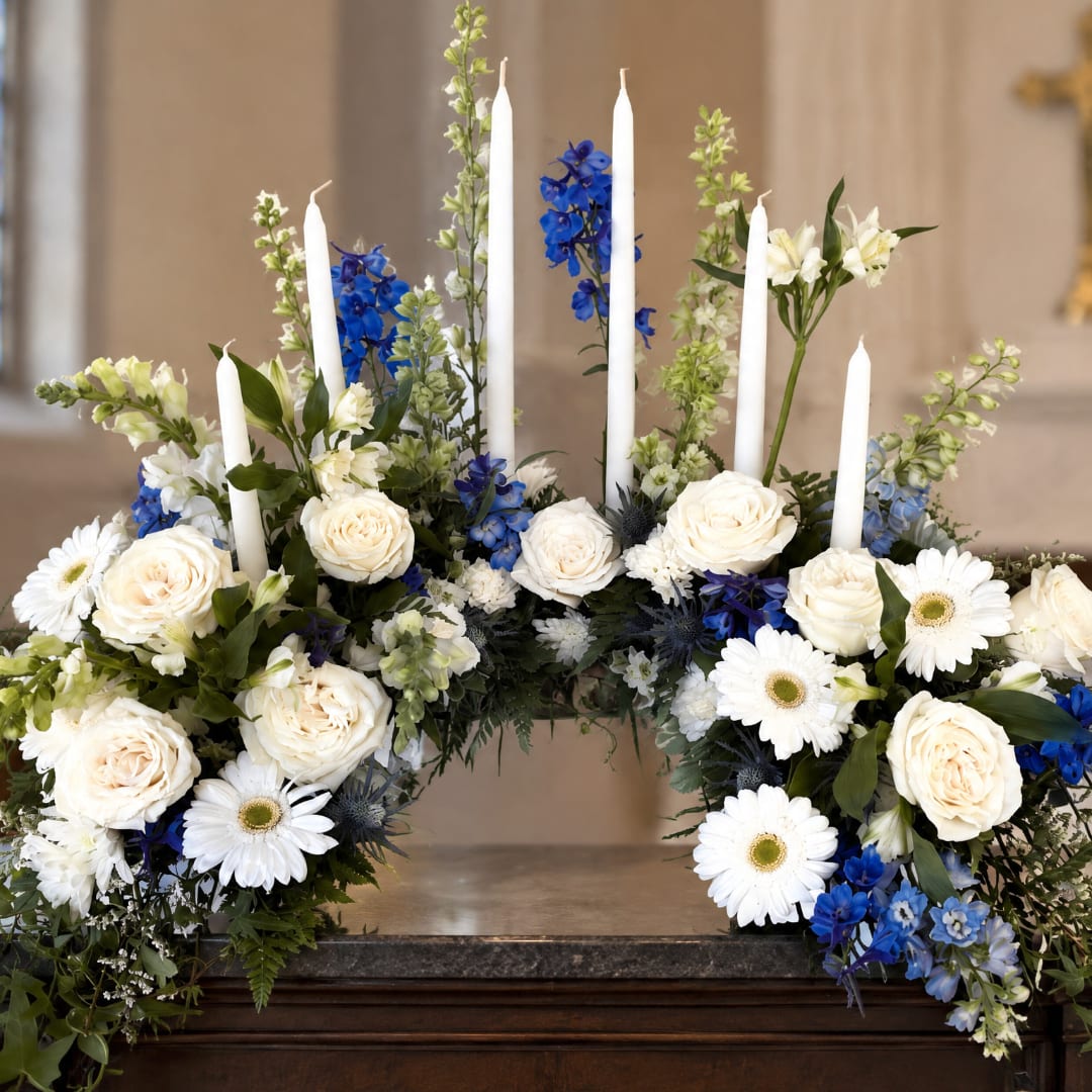 White roses and daisies arranged with blue flowers and tall white candles