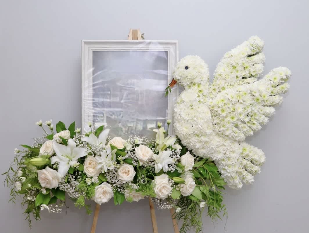White floral funeral tribute with roses, lilies, and a dove-shaped spray on an easel