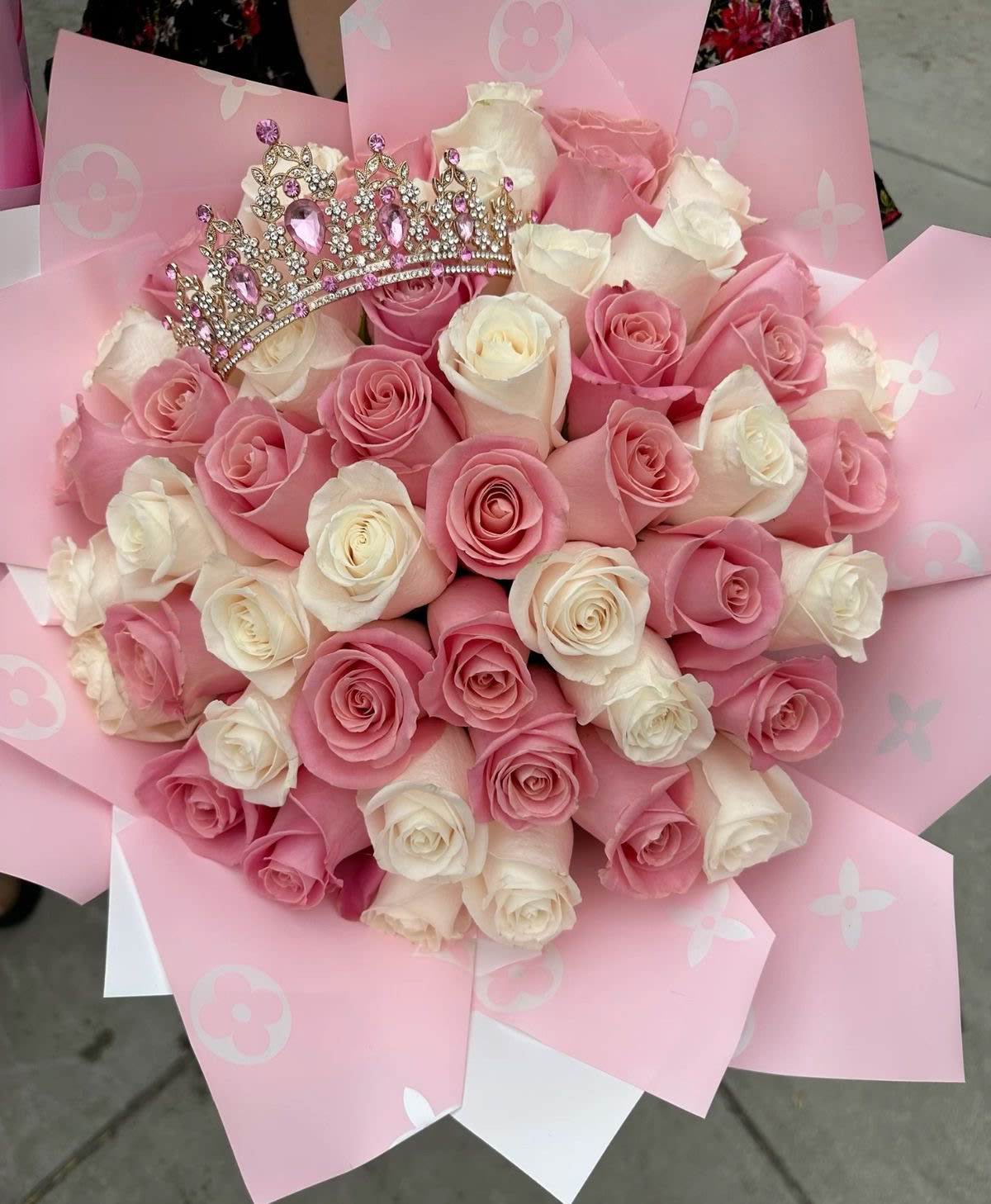 The Queen  Bouquet  - A crown of pink and creamy white roses creates an instant princess moment-perfect as a birthday bouquet, milestone celebration gift, or romantic anniversary surprise. The round, tightly clustered roses present a plush, full look that reads both elegant and playful, so the person you're celebrating will feel truly special the moment they see it.  Wrapped in layered decorative pink paper with a subtle floral pattern, the bouquet is framed by crisp, artful folds that enhance the arrangement's soft pastels. A sparkling jeweled tiara rests atop the blooms, adding a festive, youthful accent that turns this into more than flowers-it's a keepsake-ready presentation that makes the recipient feel honored and adored.  Give this to a daughter, sister, best friend, or partner when you want them to feel celebrated and seen; it's the kind of bouquet that photographs beautifully and sets a joyful tone for the day. Handle with care: keep roses in fresh water and away from direct heat or sunlight to prolong bloom life.