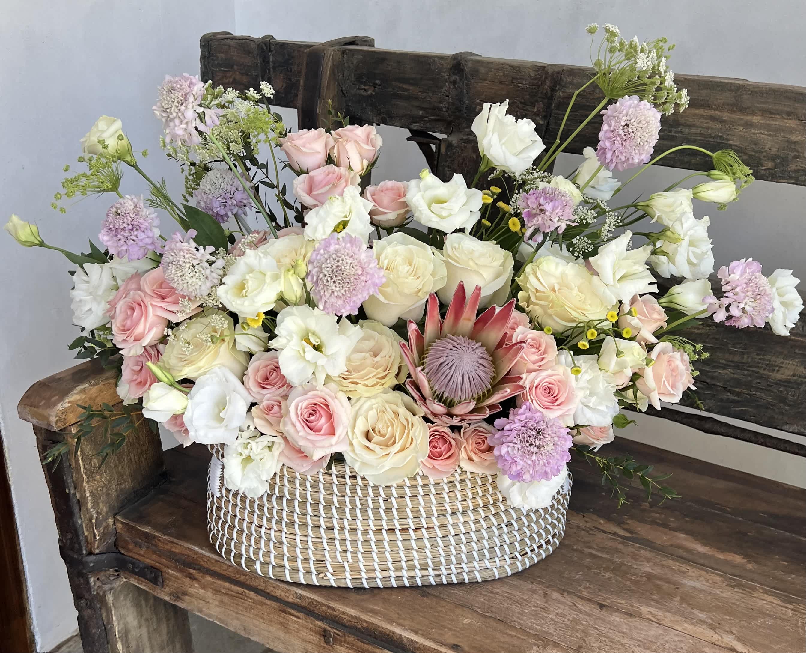 Protea Flower Basket - This flower basket features a striking protea with cream and soft pink petals, surrounded by lilac scabiosa and lush greenery.  Note: In the event that the basket featured in our picture is no longer available, we will replace it with the closest similar option, ensuring the same or better quality and size. Our goal is to maintain the beautiful presentation of your arrangement.