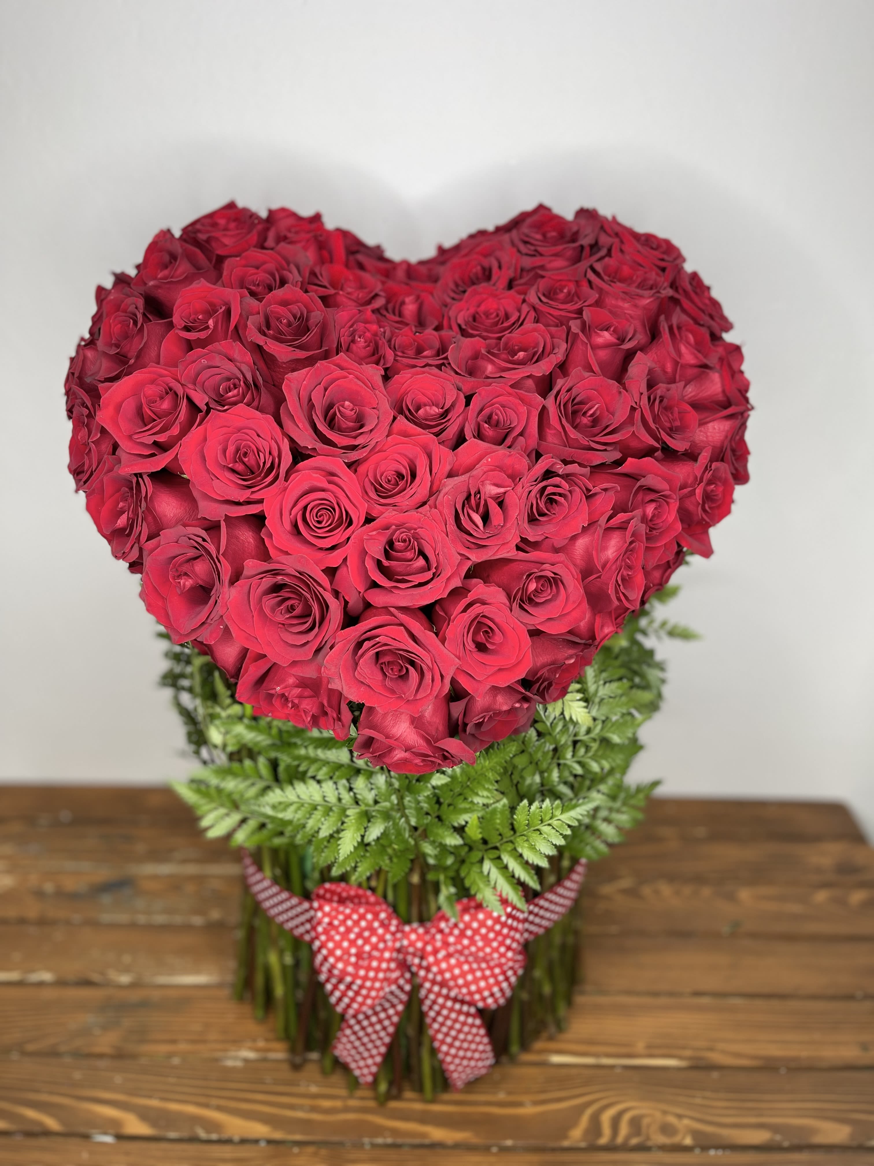 Corazón de Rosas  - An elegant heart-shaped arrangement crafted with dozens of lush red or any cloro roses of your preference, symbolizing passion and romance. The roses sit gracefully above a bed of fresh greenery, all showcased on a wooden stand that adds warmth and sophistication. Finished with a charming red polka-dot bow, this statement piece is perfect for grand romantic gestures, anniversaries, and Valentine’s Day displays.
