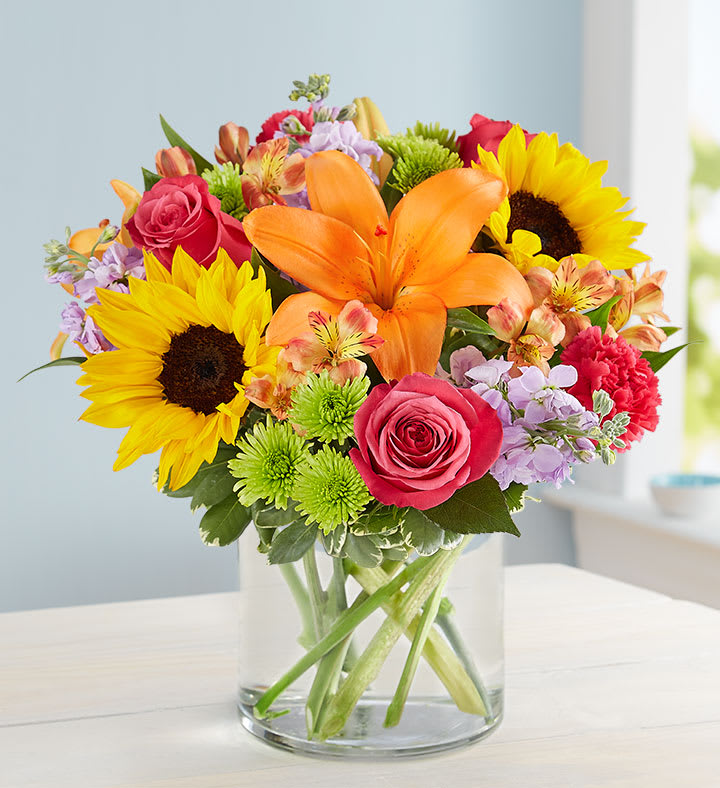 Floral Embrace - Like a warm embrace, our vibrant bouquet delivers your sentiments to someone special. A rich gathering of yellow and orange blooms, with pops of bright pink and purple, it's more than a gift - it's a way to express how you feel inside. All-around arrangement of hot pink roses and carnations; orange Asiatic lilies and Peruvian lilies (alstroemeria); yellow sunflowers or similar blooms; lavender stock; purple monte casino; green Athos poms; accented with assorted greenery Artistically designed in a glass vase Large arrangement does not include carnations