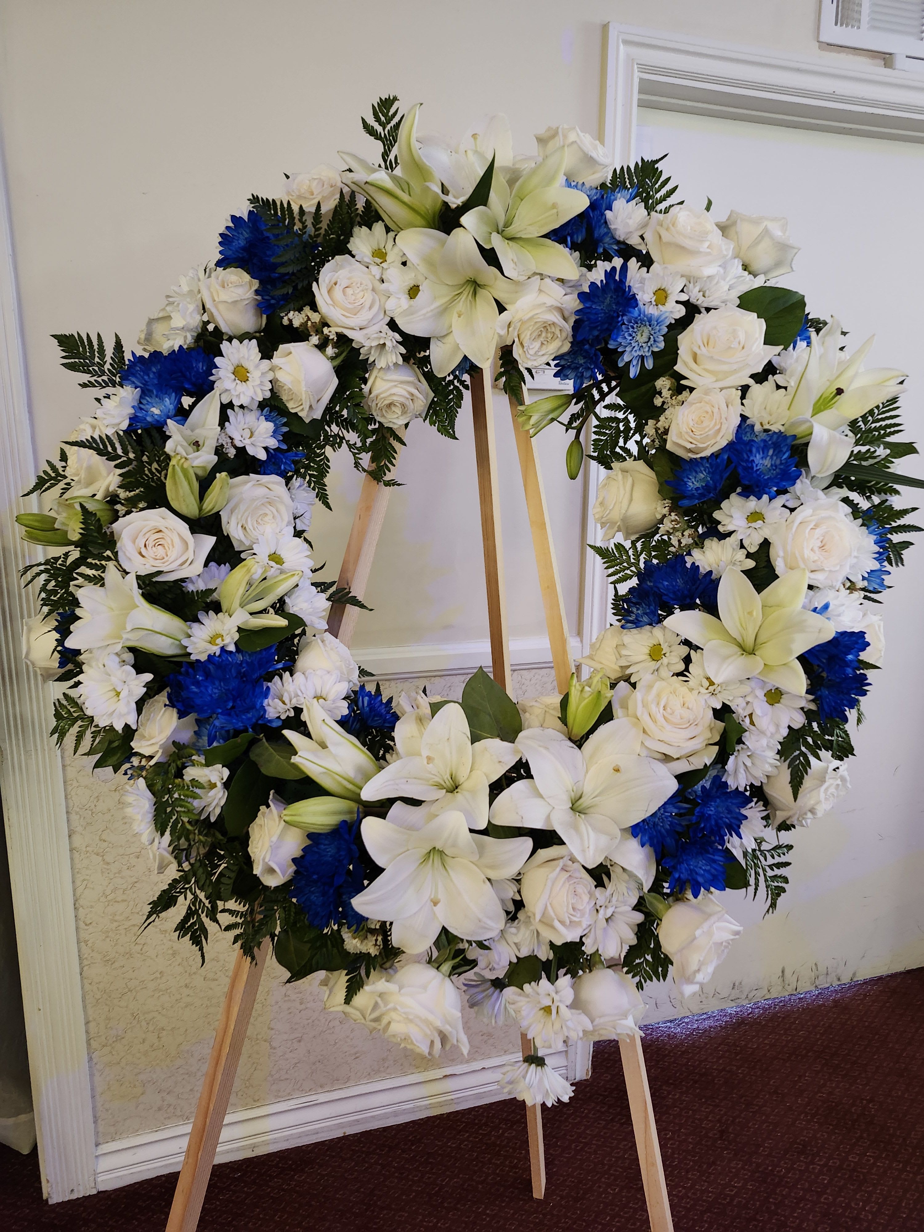 Grand Wreath Blue/White  - Large blue and white sympathy wreath. Perfect for a male's services. It is designed with roses, lilies, daisies, cushion pompoms and wax flowers. The banner is sold separately by searching "Sympathy Banner"
