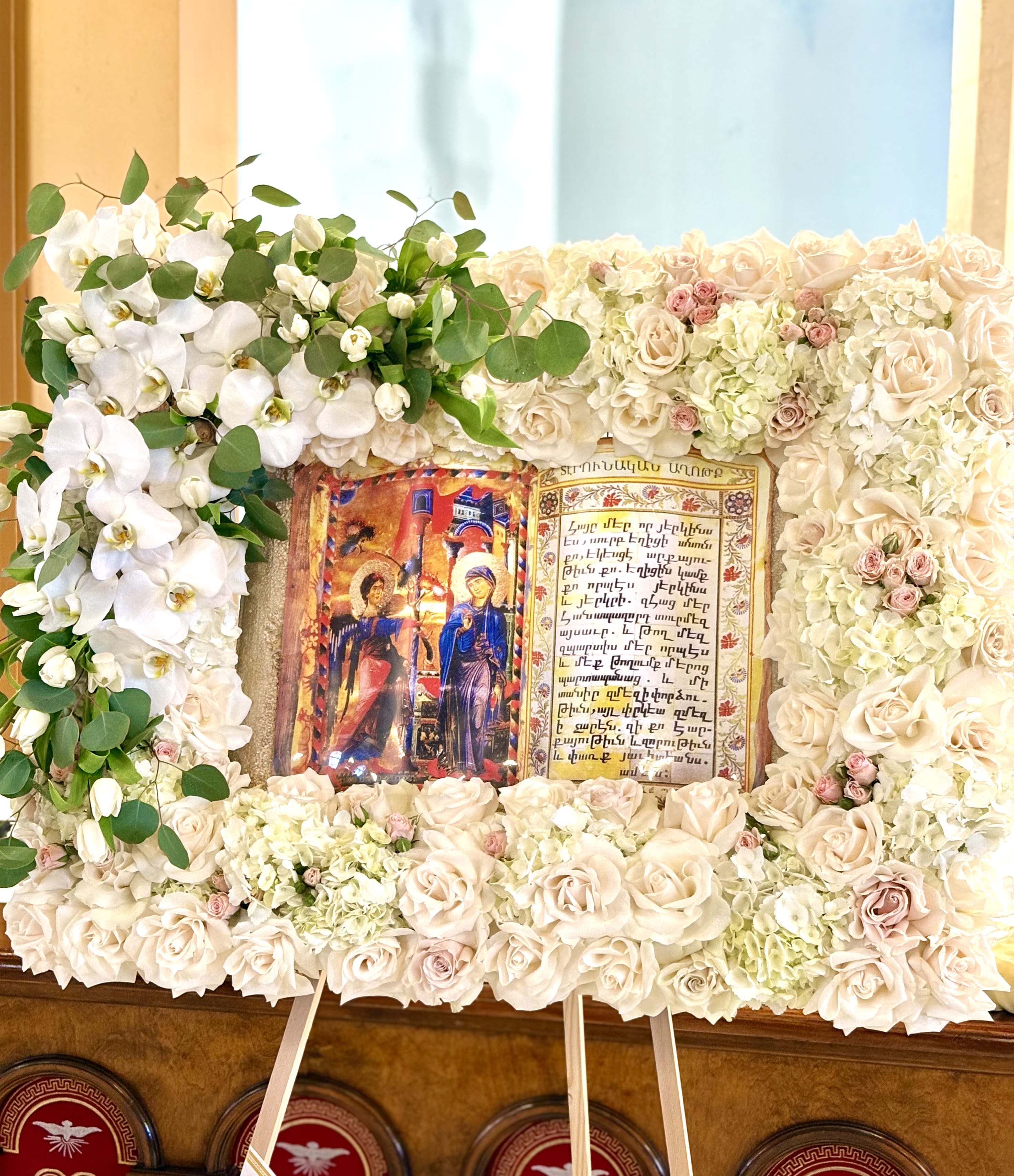 "The Bible" By VLM Flowers - A reverent presentation piece centered on a religious icon, this rectangular floral frame is designed as a ceremonial flower arrangement for memorials, funerals, or church dedications. The composition reads like a floral "book" around the central text panel, so the person you're honoring will be framed with a quiet, respectful presence that suits formal services and moments of remembrance.  Draping white Phalaenopsis orchids sweep down the left side while pale cream roses and soft pink spray roses build the top, bottom, and right edges into a dense, symmetrical border. Soft green foliage and touches of hydrangea fill and support the frame, giving a full, textured backdrop that looks substantial from a distance. The arrangement comes finished on a display easel with the included religious icon and central panel, creating a polished, dignified tribute.  Made to be seen at services or placed in a reception area, this piece communicates care and solemn respect-ideal for honoring faith, life, and memory. Ask about sizing or delivery options to ensure it fits the service space; flowers arrive professionally mounted and need only the easel placed where the ceremony will best receive the tribute.