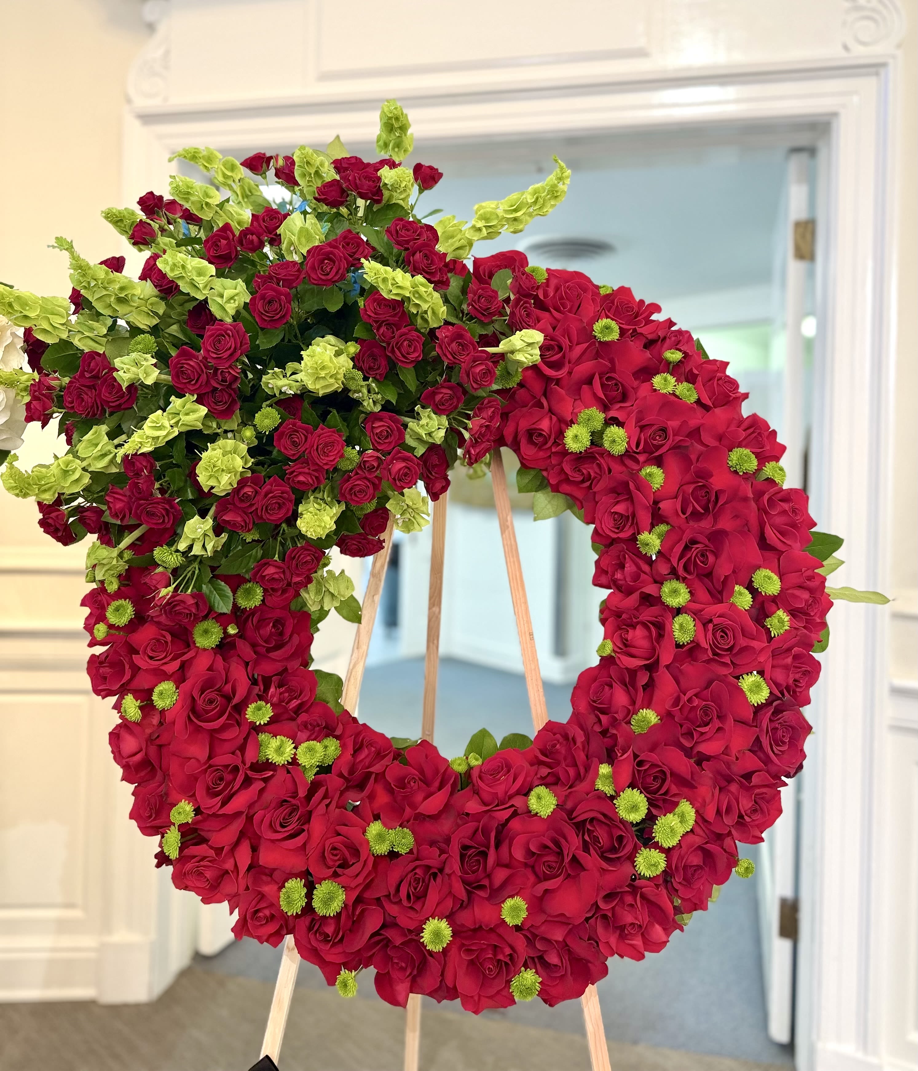 Graceful Remembrance By VLM Flowers - A large funeral wreath built from richly packed red roses creates an immediate, heartfelt impression-perfect as a sympathy tribute or a formal expression of love for a memorial service. This floral arrangement for remembrance conveys deep affection and solemn respect; the person you're honoring will be remembered with a striking symbol of devotion that reads clearly from the front of the room.  Densely arranged red roses form the core of the design, accented with bright green button chrysanthemums and lime-green clusters that lift the composition and add fresh contrast. Mounted on a sturdy wooden easel, the wreath stands tall for easy display and photographs; the bold red and green palette gives the piece both romantic warmth and dignified presence. This large-scale arrangement is intended for services and memorials where a powerful visual tribute is appropriate.  