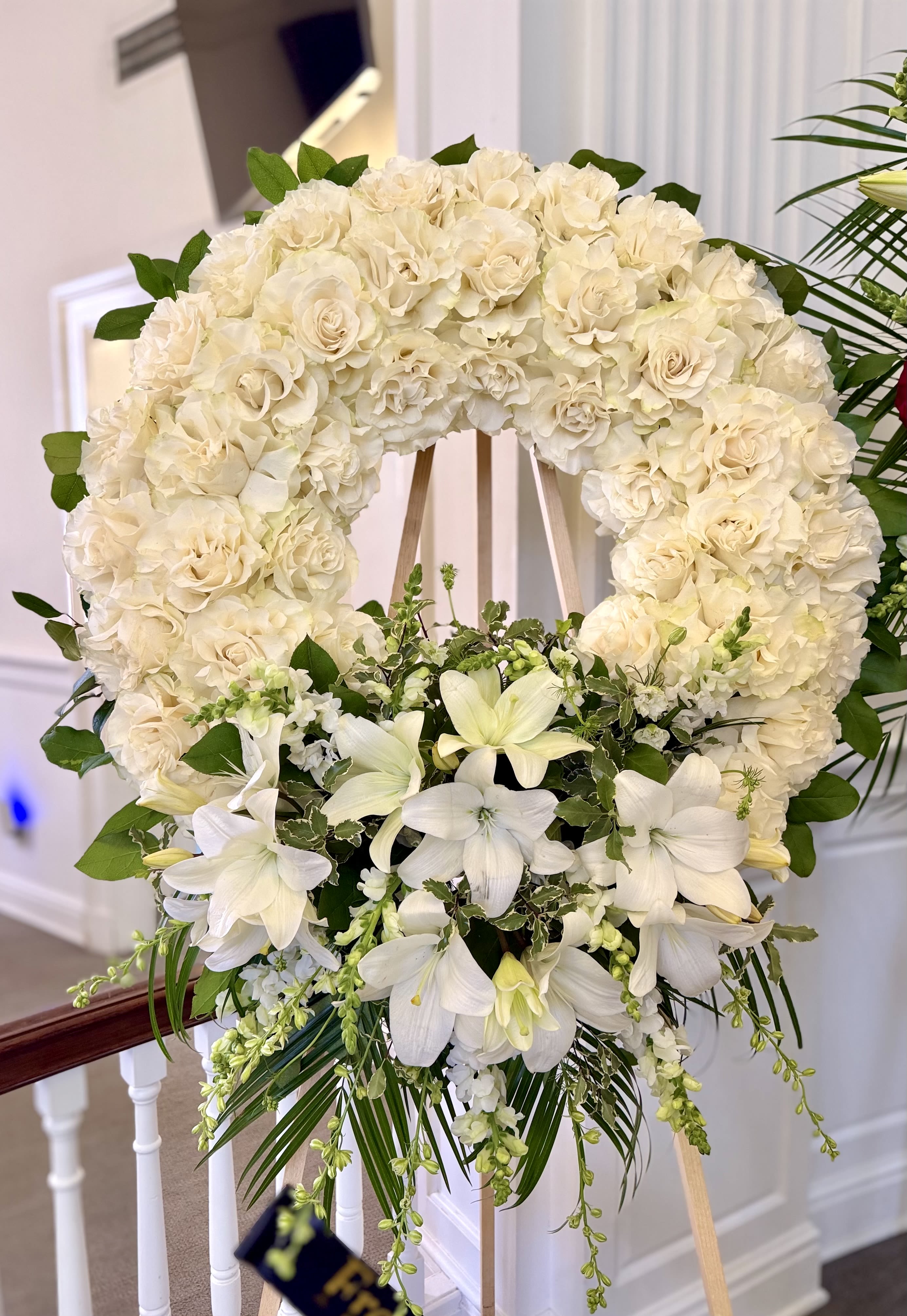 Graceful Remembrance Wreath By VLM Flowers - A large circular funeral or memorial wreath crafted in soft whites and gentle greens-designed to offer a serene, dignified tribute for a memorial service or sympathy display. The arrangement's creamy white roses and  blooms form a dense, elegant ring while a clustered spray of white lilies at the base adds a graceful, cascading focal point that draws the eye and soothes the heart.  