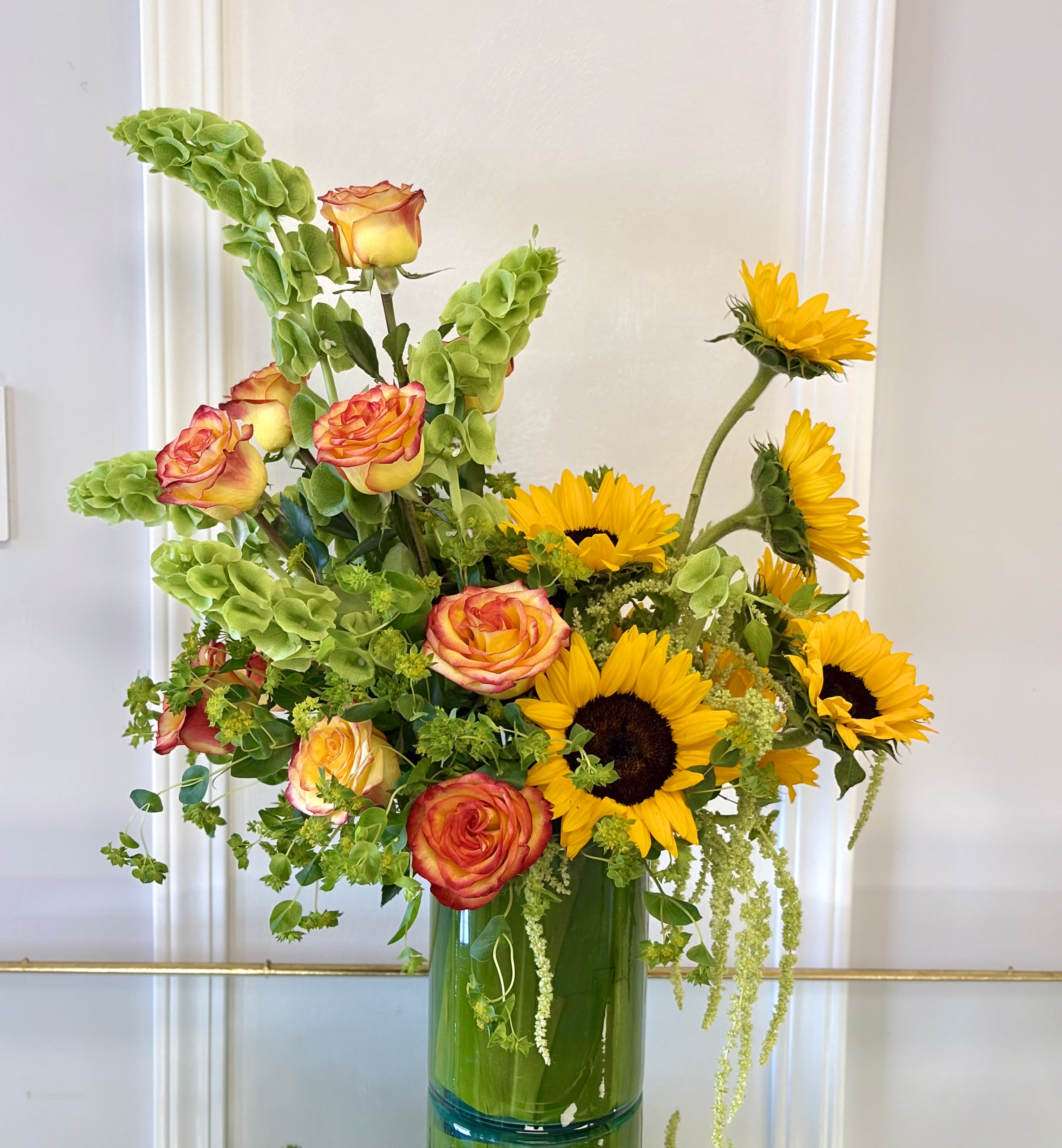 Sunlit Elegance By VLM Flowers - A striking arrangement featuring radiant sunflowers, premium bi-color roses, vibrant Bells of Ireland, and lush seasonal greenery. Designed with height, texture, and natural movement, this bouquet brings warmth and sophistication to any celebration.