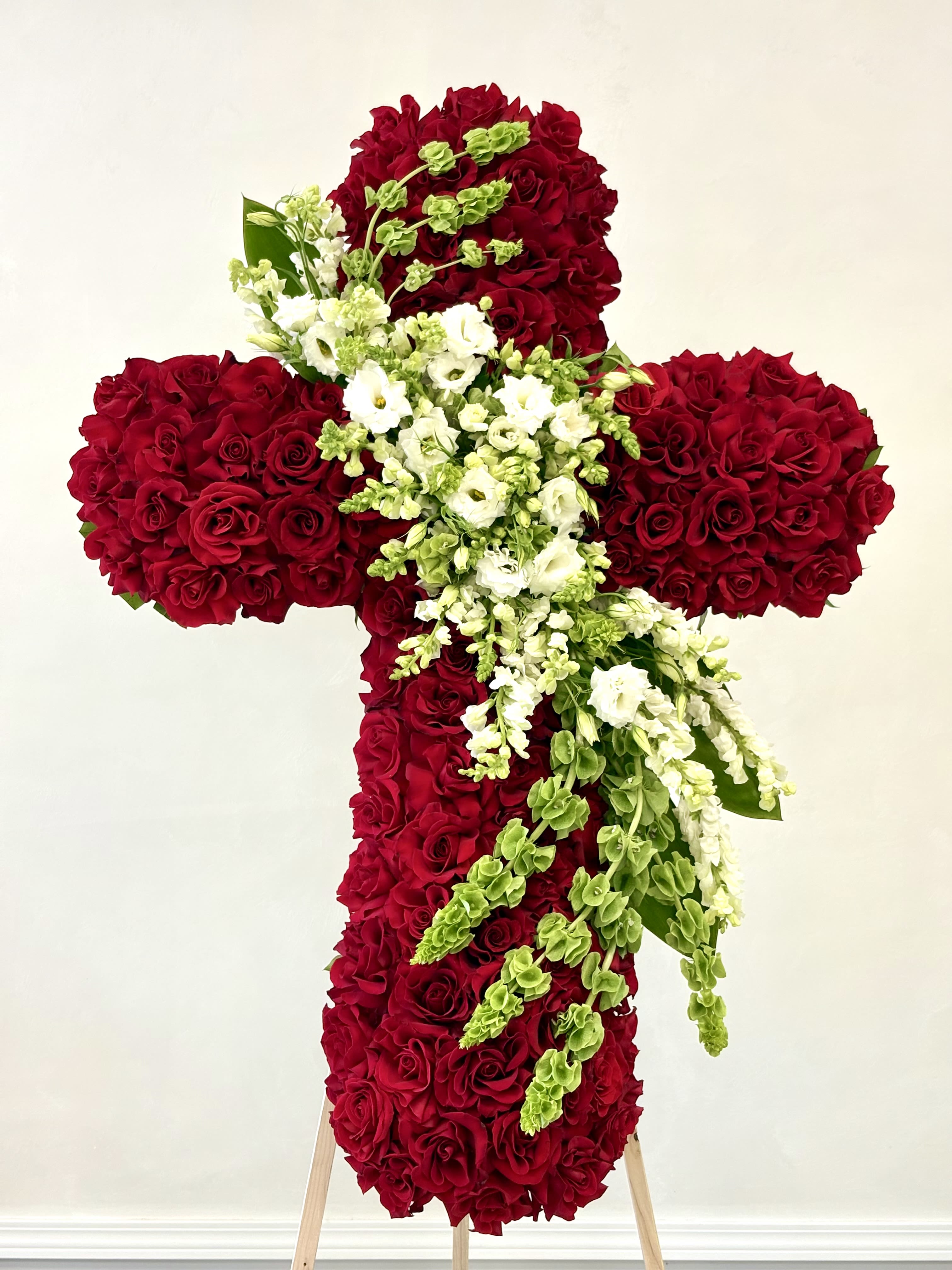 Forever in Our Hearts Cross By VLM Flowers - An exquisite standing cross of lush red roses accented with cascading white stock, lisianthus, and Bells of Ireland. This elegant tribute expresses profound sympathy, enduring love, and the promise of everlasting peace.