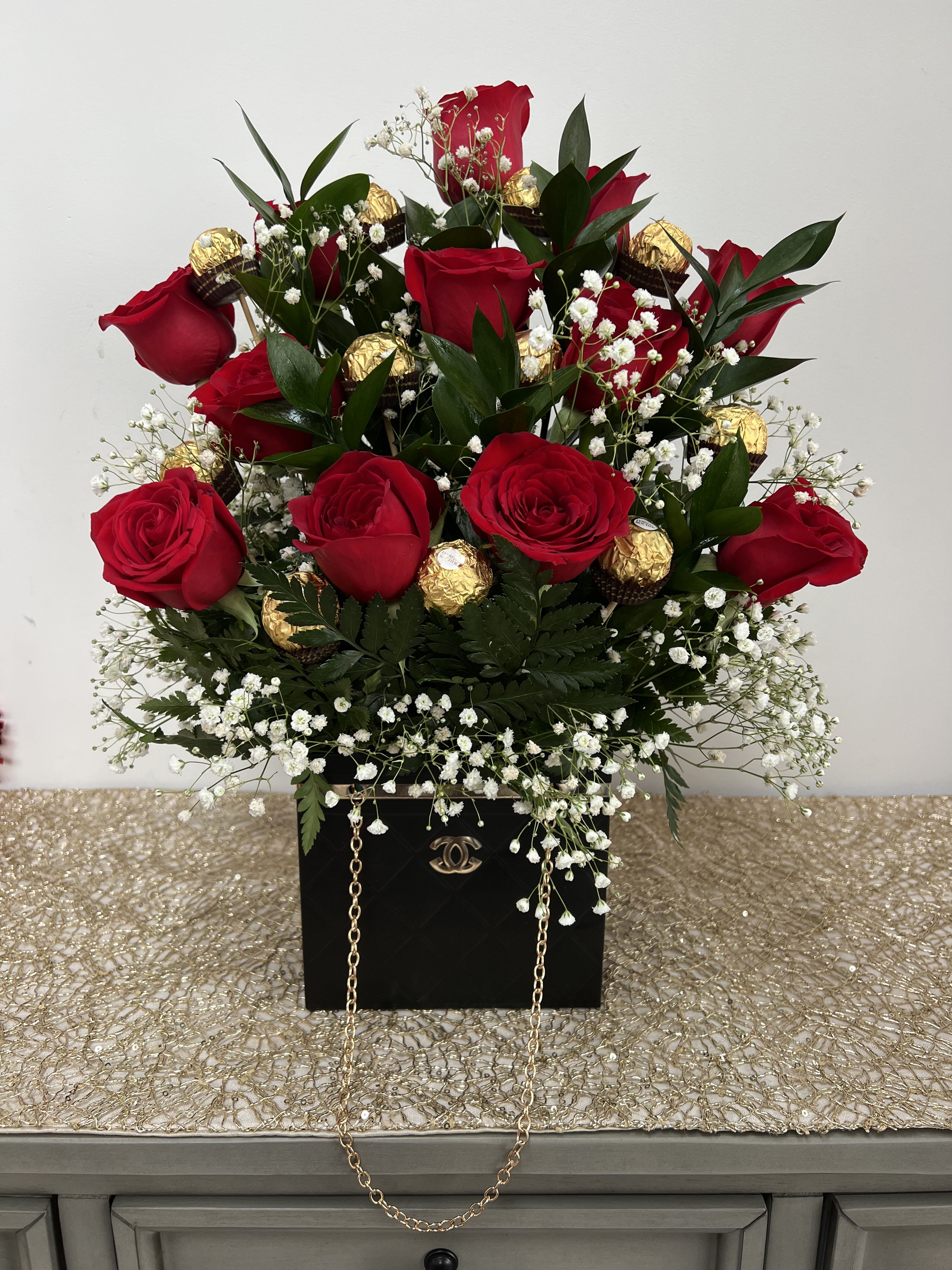 Rosé Noir Delight - A stunning arrangement that blends timeless romance with a touch of luxury. This elegant bouquet features rich red roses nestled among fresh greenery and delicate baby’s breath, beautifully accented with premium gold-wrapped chocolates. Designed in a chic black keepsake box with a gold chain detail, it makes a bold and sophisticated statement. Perfect for anniversaries, birthdays, Valentine’s Day, or any special occasion, this arrangement delivers both beauty and indulgence in one unforgettable gift.
