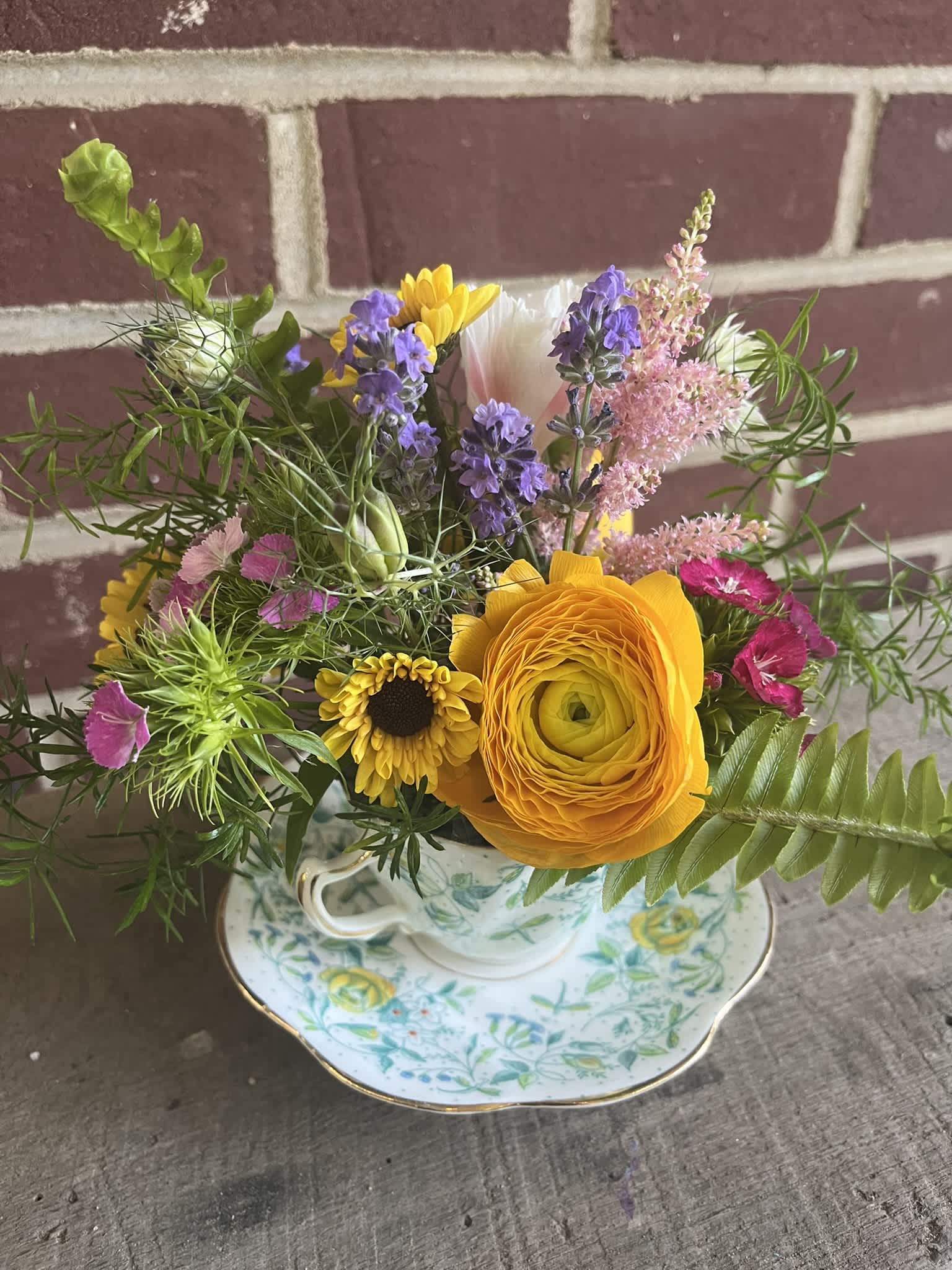 Mixed bouquet in a floral teacup and saucer with yellow, purple, pink, and white blooms