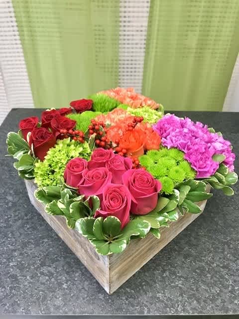 Square box arrangement of pink and red roses, green mums, orange carnations, and pink blooms in color-blocked sections