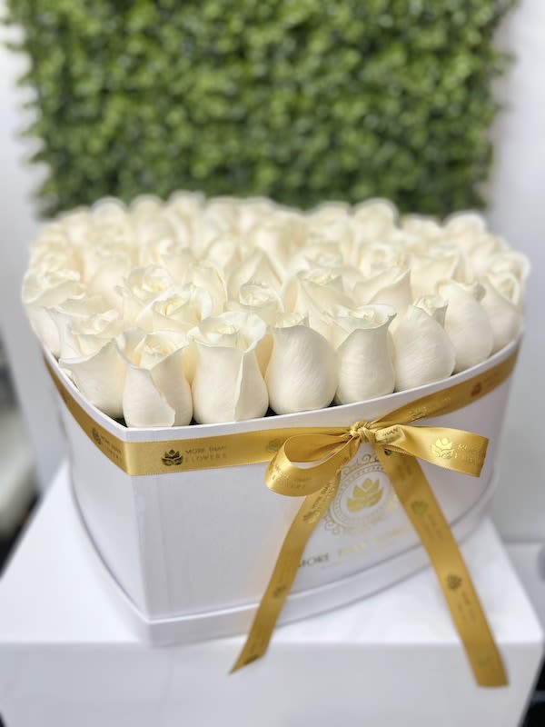 Heart-shaped white box filled with tightly packed ivory roses and tied with a gold satin ribbon.