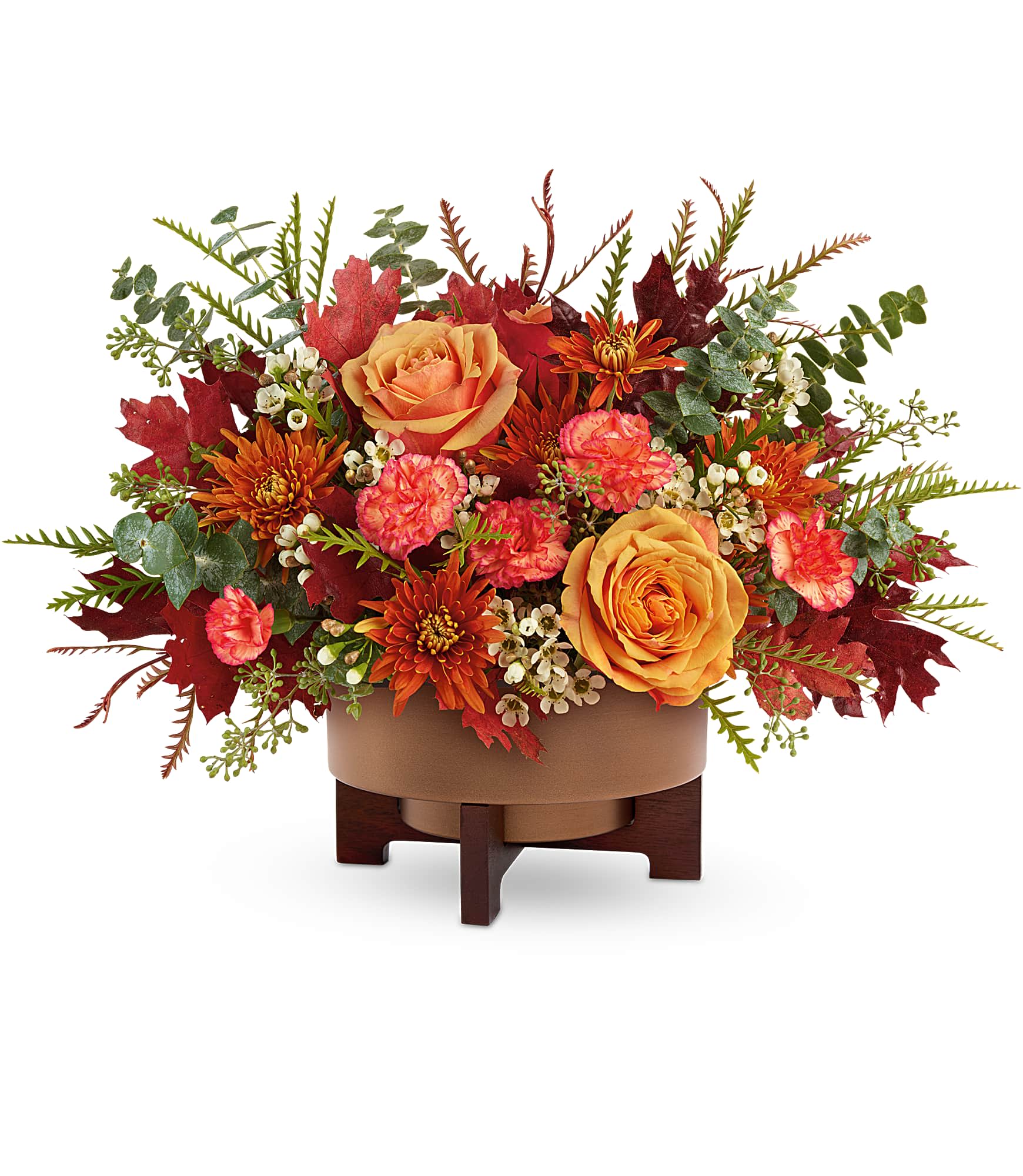 Modern Copper Centerpiece - Bring warm, autumnal elegance into any space with this stunning floral arrangement. Featuring a rich blend of sunset-orange roses, vibrant coral carnations, and rust-toned mums, this centerpiece captures the cozy beauty of the fall season. Accented with white wax flowers, fresh eucalyptus, and seasonal foliage in deep red and copper hues, it creates a lush, layered look full of texture and color.  Artfully arranged in a modern copper-toned bowl elevated on a dark wooden stand, this design offers a perfect balance of contemporary style and natural charm. Ideal for holiday tables, home décor, or seasonal gifting, it adds warmth and sophistication to any setting.