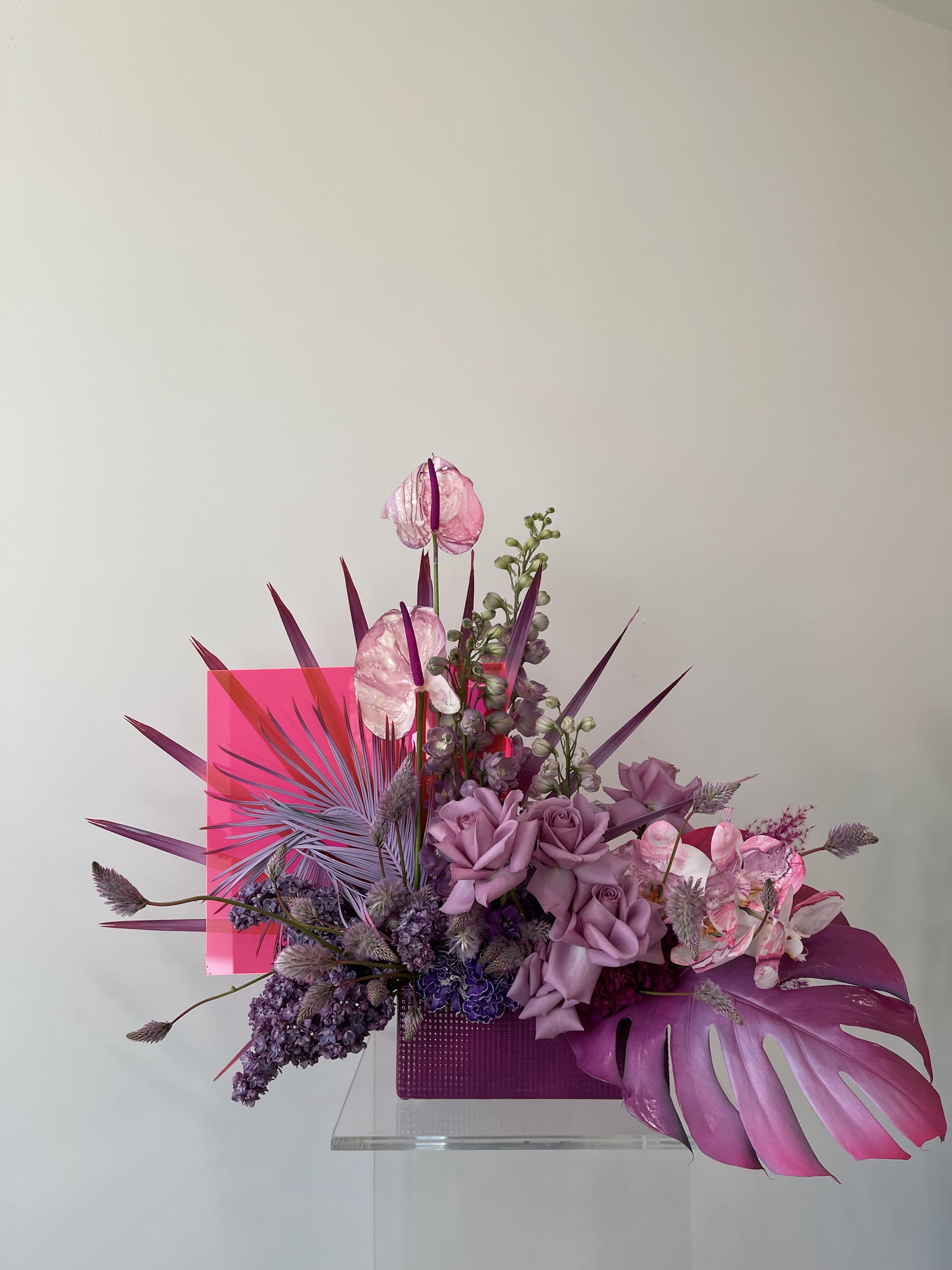 Purple floral arrangement with roses, orchids, and tropical leaves in a pink container