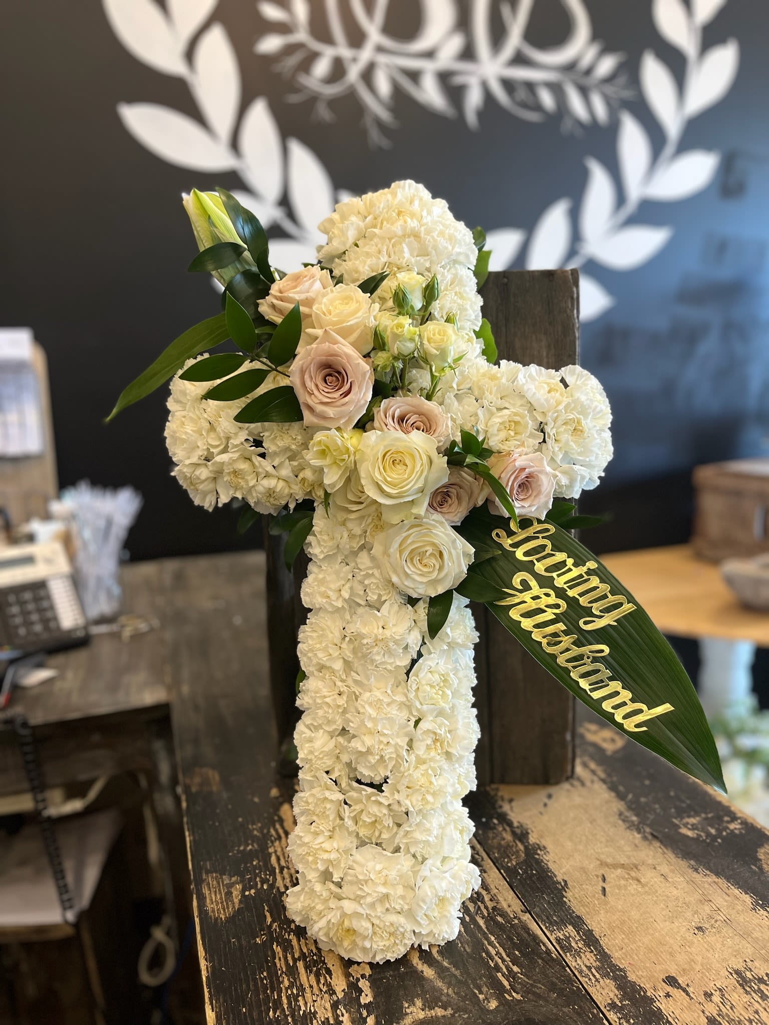 White floral cross arrangement with pale roses and a gold "loving husband" ribbon