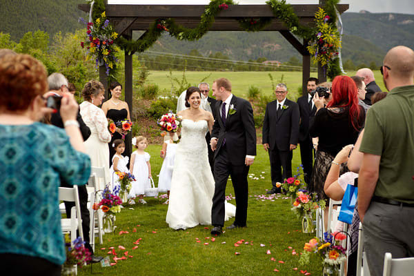 Bride and groom walking down an outdoor wedding aisle with floral arch and guests