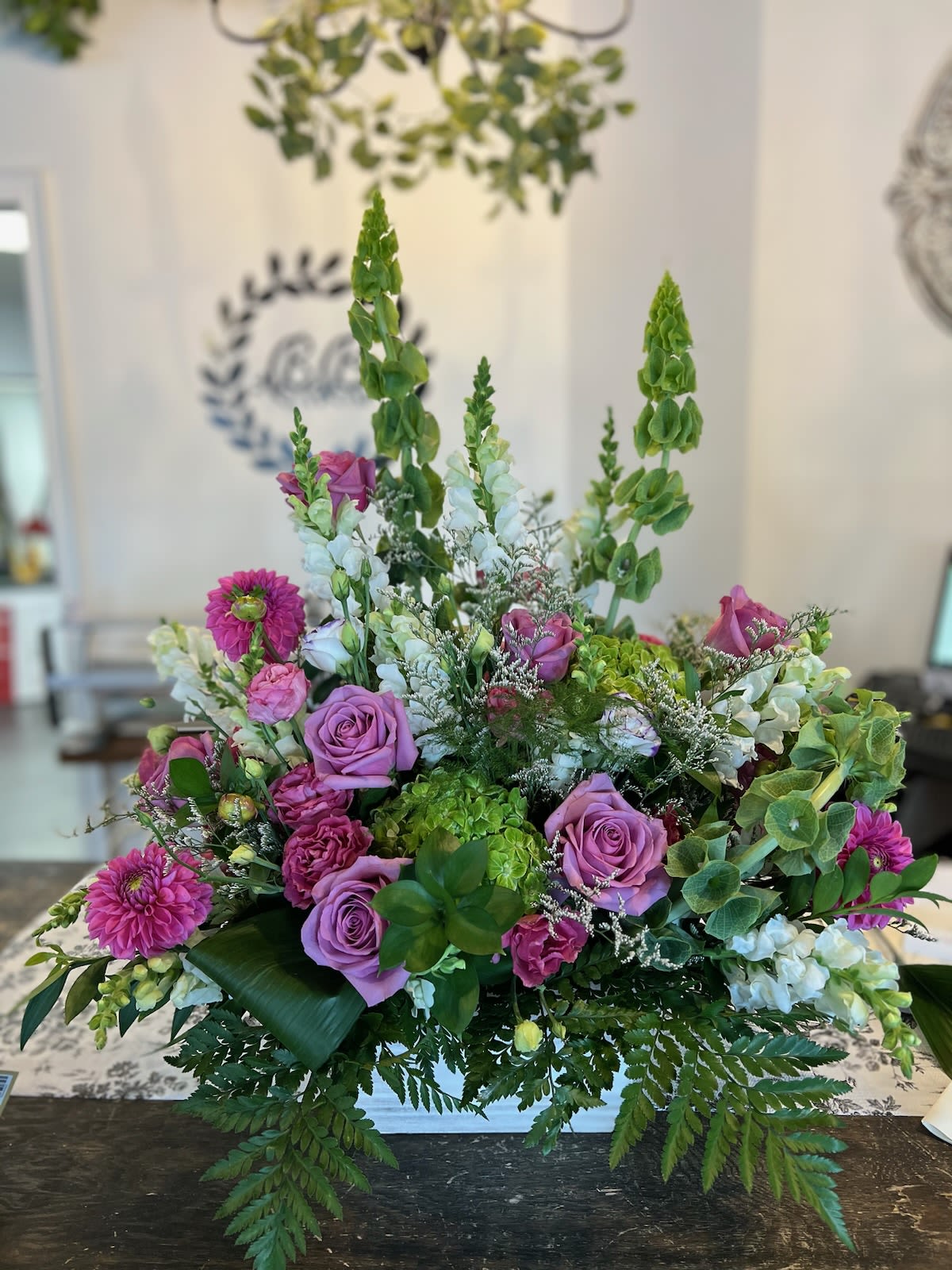 Lush arrangement of purple roses, pink blooms, white flowers, and green Bells of Ireland in a low white container.