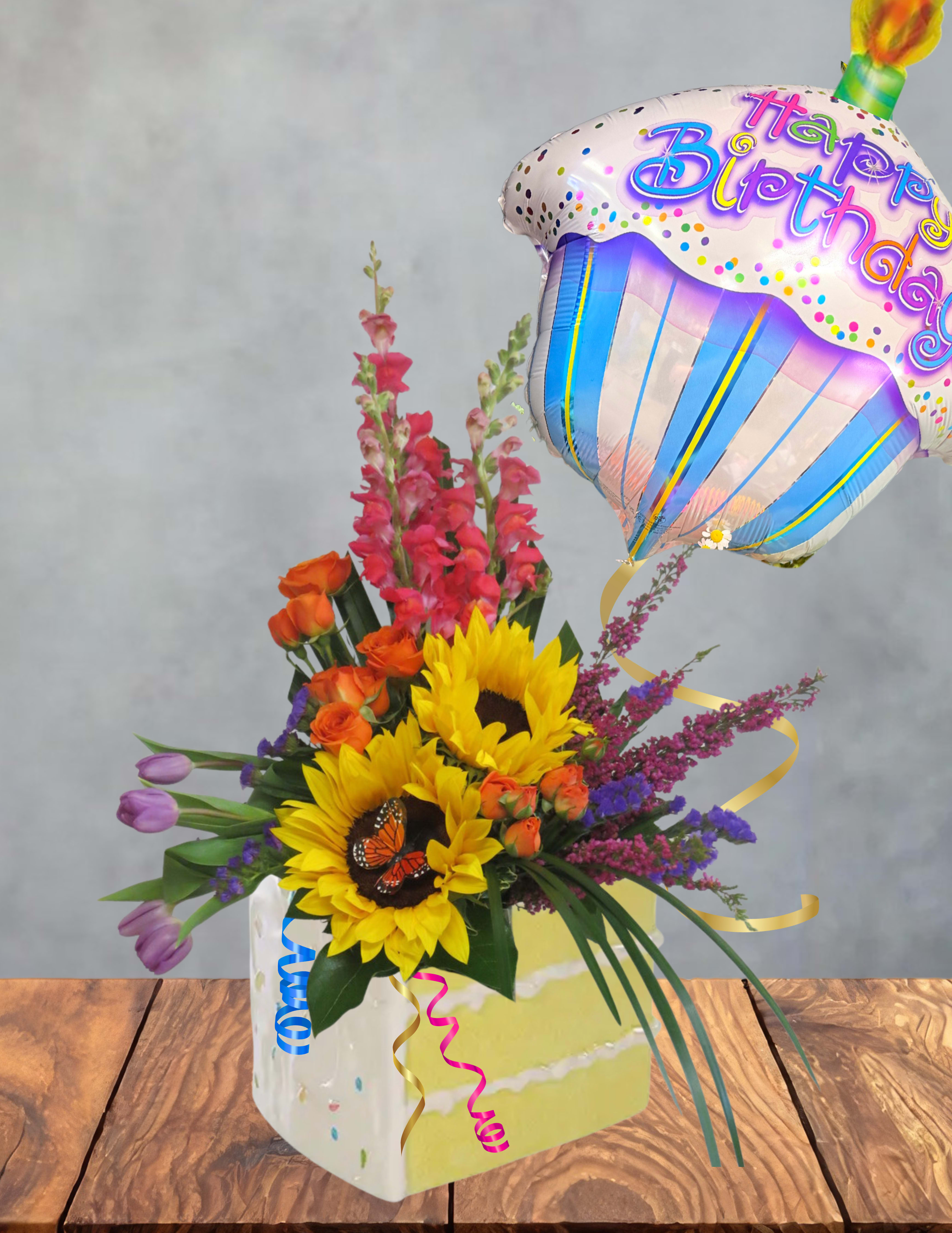 Confetti cupcake  - Bright sunflowers steal the show in this birthday bouquet, paired with orange roses, purple tulips, and tall pink snapdragon spikes to create a Confetti Cupcake arrangement that feels joyful and celebratory. Designed as a birthday gift arrangement, the mix of warm yellows and oranges with cool purples and pinks gives the person you're thinking of an instant burst of party energy-the kind of floral surprise that says "celebrate you" the moment they see it. A large birthday balloon rising from the design adds playful height and a festive focal point.  Flowers are artfully layered in a decorative gift box, tied with a coordinating ribbon for a polished, ready-to-give presentation. The composition pairs the bold, textured faces of sunflowers with the soft romantic curves of roses and tulips and the vertical interest of snapdragons, creating depth and movement that reads beautifully from every angle. Ideal for birthdays, milestone celebrations, or to brighten someone's day, this arrangement arrives complete with balloon and gift-box styling for easy gifting and display.