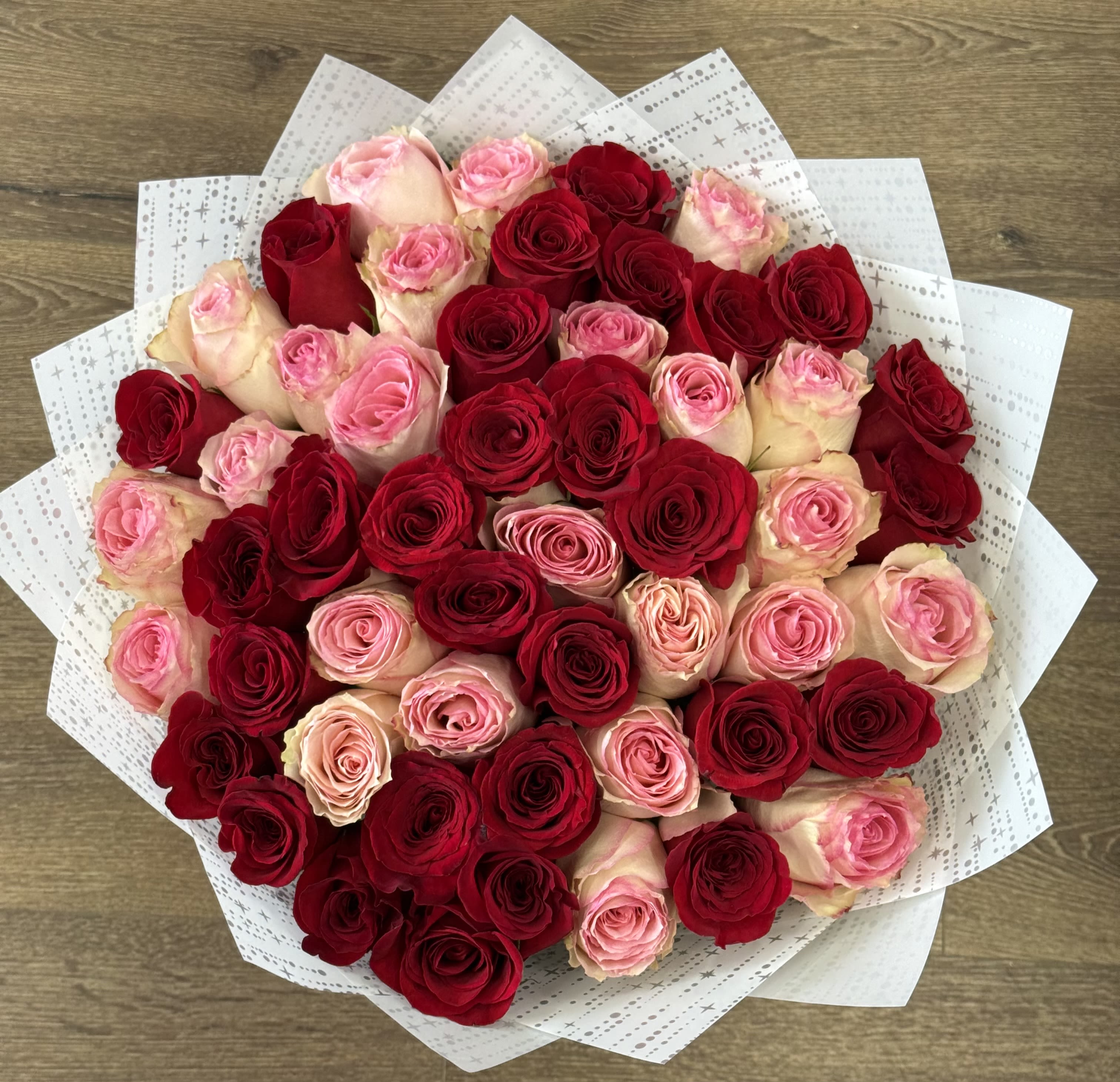 Strawberry Sundae Bouquet - The Strawberry Sundae is a hand-tied arrangement featuring a dense, rounded mound of premium Red and Pink Roses. The design utilizes a mix of deep red and bright pink tones to create a high-contrast floral display. It is finished in a structured White Korean Paper wrap with a semi-transparent border, which is designed to emphasize the vivid saturation of the rose petals.