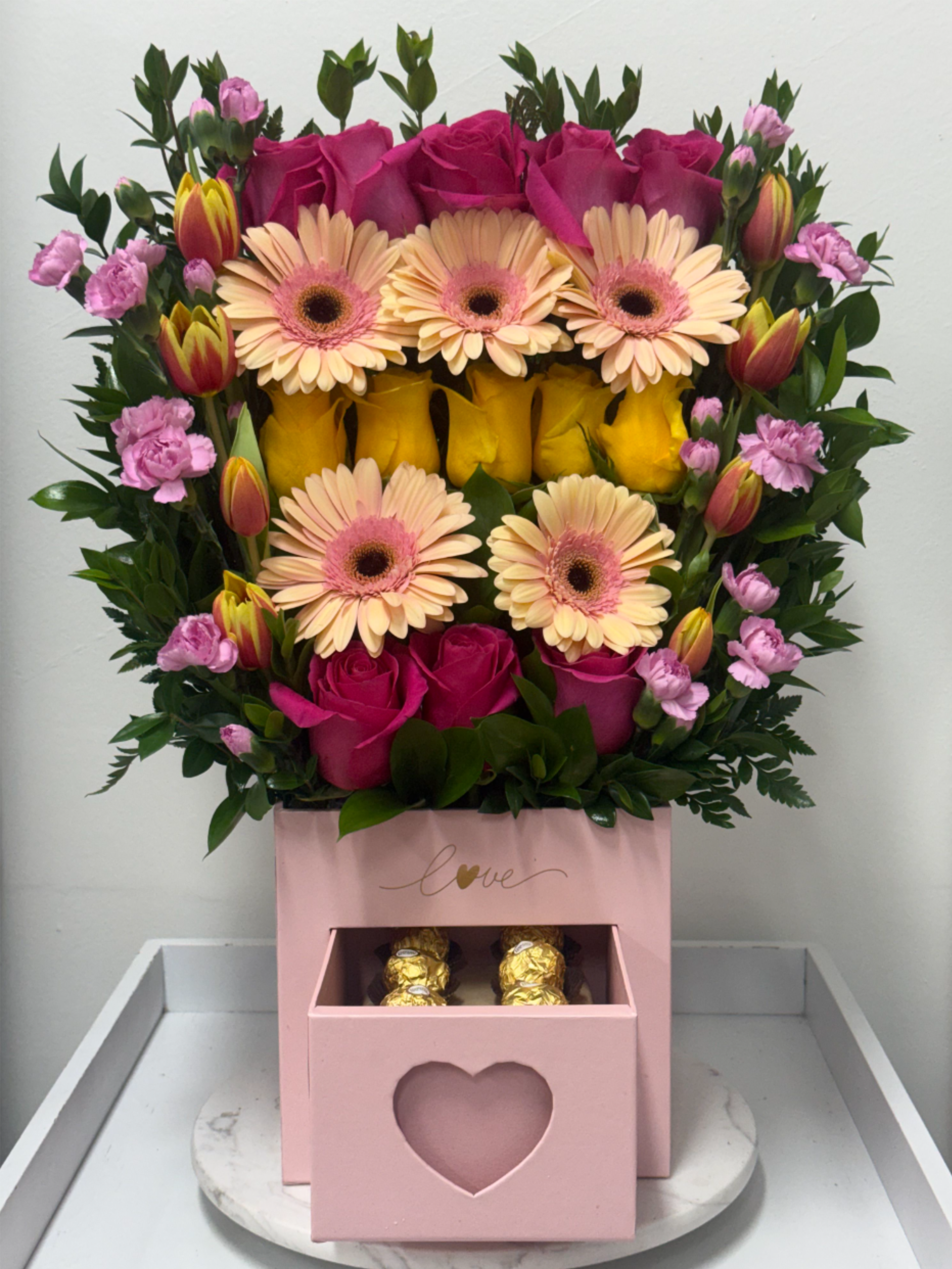 Pink Eye Blossom Box - The Pink Eye Blossom Box features a multi-layered arrangement of hot pink and yellow roses, pink gerbera daisies with contrasting dark centers, bi-color red and yellow tulips, and miniature pink carnations. This design is framed with assorted green foliage and presented in a pink box with an integrated bottom drawer containing gold-wrapped chocolates.