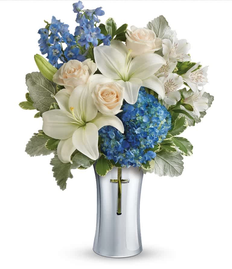 Skies Of Remembrance - As hopeful as a bright blue sky, this beautiful arrangement of lush blue hydrangea and fragrant white lilies is a heartfelt symbol of peace and beauty. Its reverent, silver-finished ceramic vase with cross cut-out will remain a guiding light to your loved ones for years to come. This majestic bouquet includes blue hydrangea, white asiatic lilies, white alstroemeria, light blue delphinium, dusty miller and variegated pittosporum. Delivered in a Shining Cross vase. Approximately 14 1/2" W x 18 3/4" H