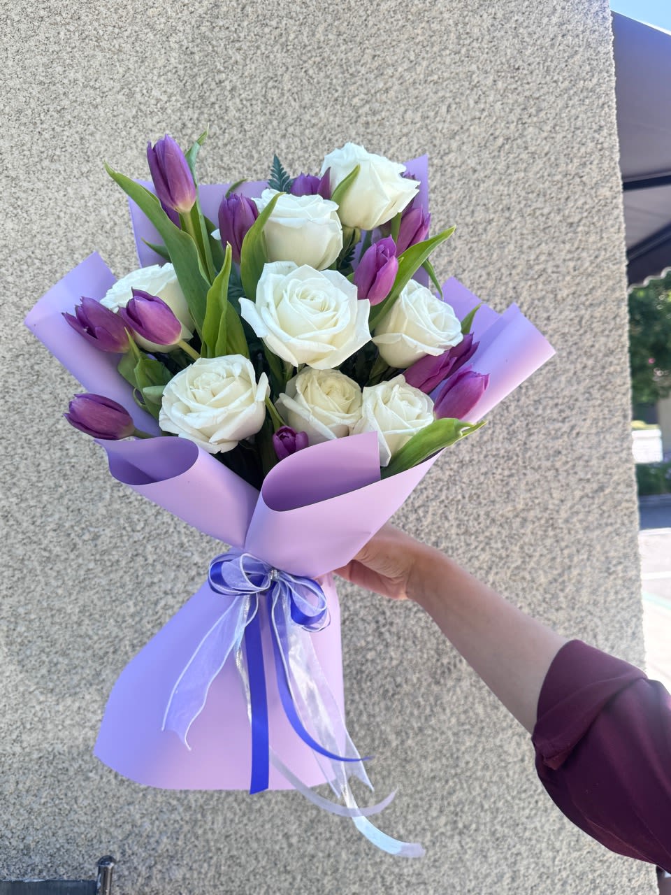 Whites Roses with Purple Tulips  - White roses and purple tulips are arranged in a rounded hand-tied bouquet that reads both romantic and fresh - a perfect birthday bouquet or anniversary gift to make someone feel cherished. The contrast of crisp white roses against deep violet tulips creates an elegant springtime palette that feels thoughtful without being overdone.  Wrapped in soft lavender paper and finished with a blue ribbon, the bouquet's pastel outer tones highlight the richer flower colors for a gentle, polished presentation. Textural interest comes from the smooth rose petals and the tulips' sleek silhouettes, making this arrangement ideal for celebrating milestones or simply telling someone you're thinking of them. Care: keep stems in fresh water, trim ends if needed, and display away from direct heat to extend vase life.