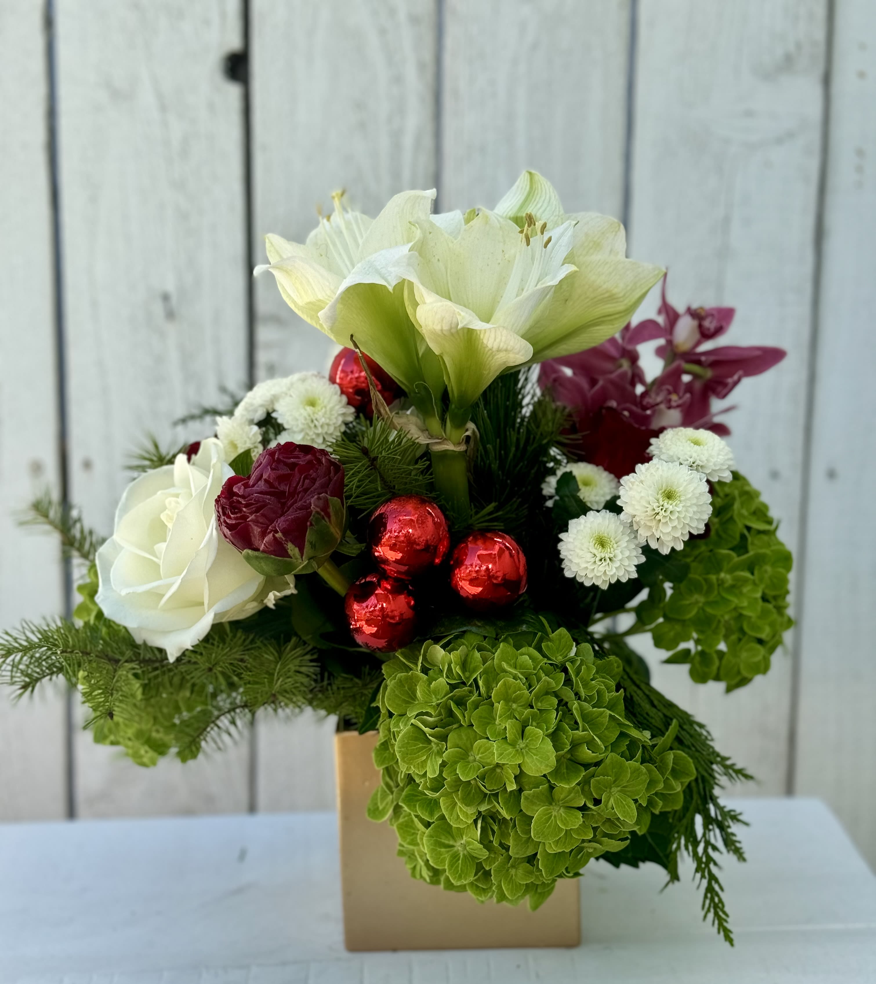 A MANHATTAN BEACH HOLIDAY - This exceptionally pretty design is so perfect for your special family and friends! Gorgeous white roses, red roses, green hydrangeas, white Amaryllis, cymbidium orchids , peonies and touches of red in decorative ornaments. It’s a nice size in a 6x5 square gold container.