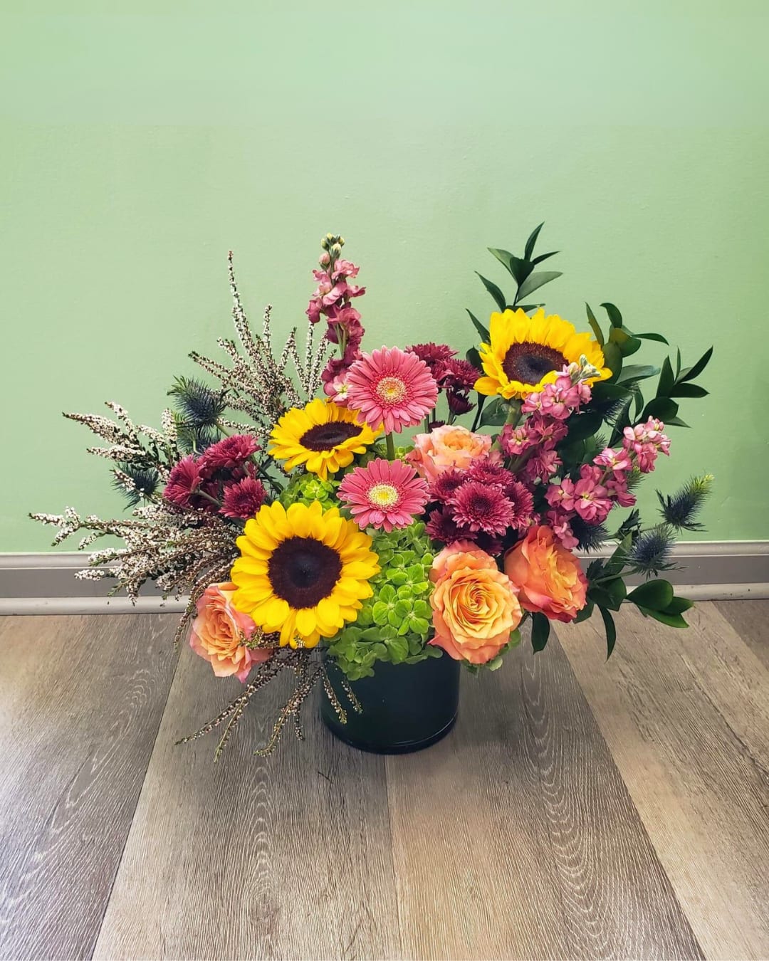 Autumn Hues - This gorgeous arrangement has stock, mini gerberas, sunflowers, calycium, mums, roses, hydrangeas, thistle, and greens in a clear cylinder vase, 