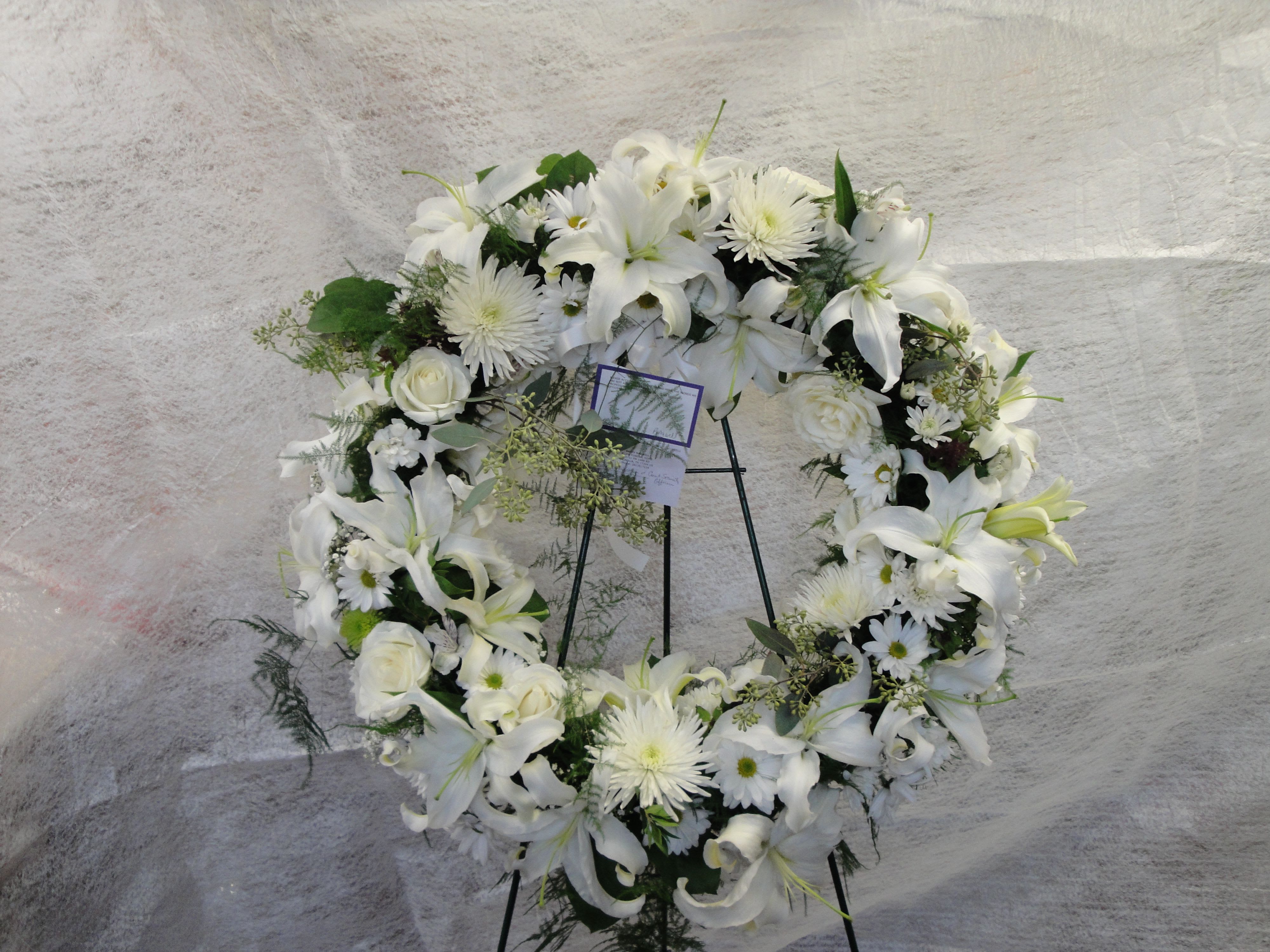 White floral wreath on a black stand with lilies and daisies