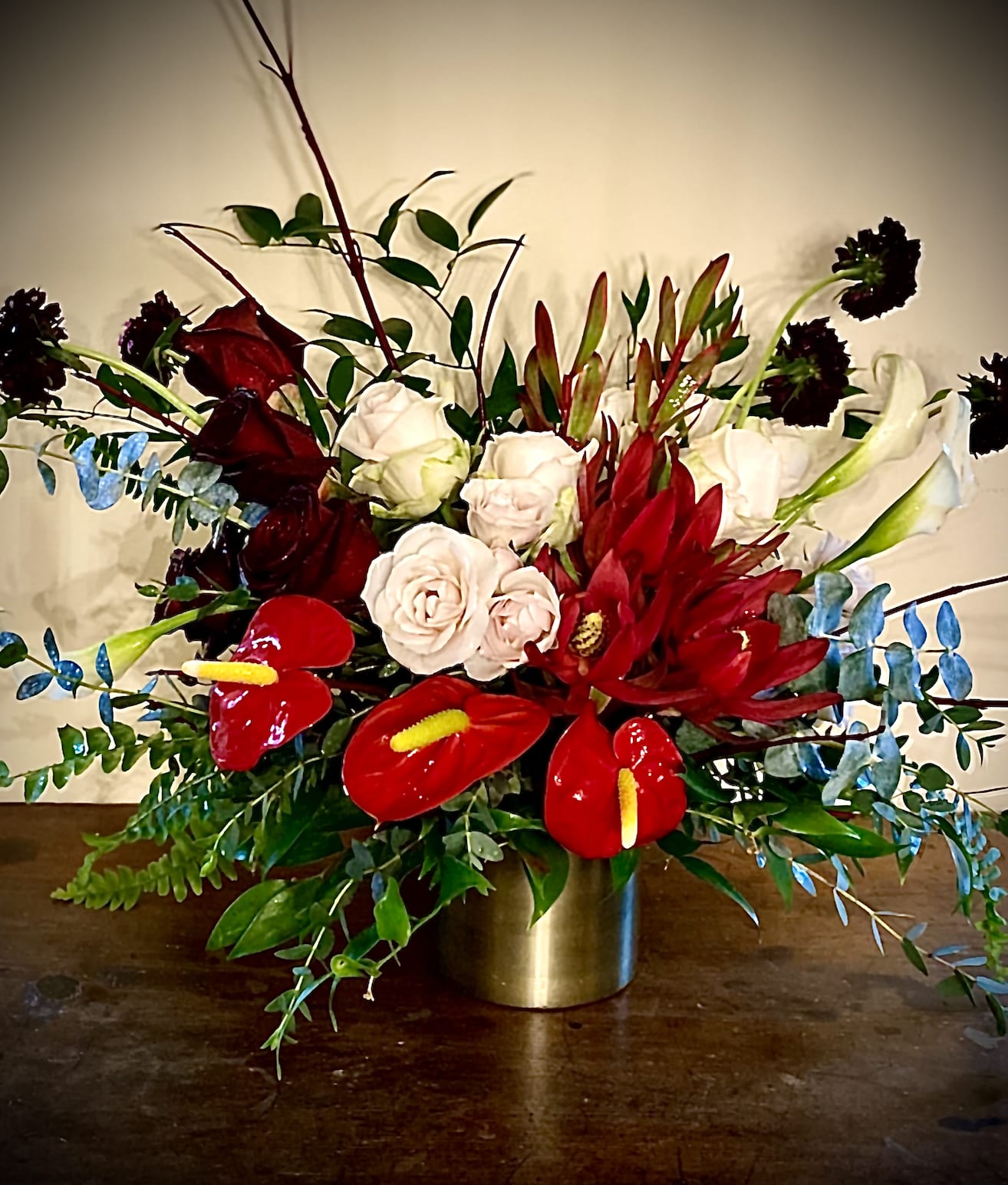 Low gold vase with red and blush roses, red tropical blooms, and white lilies in a lush arrangement.