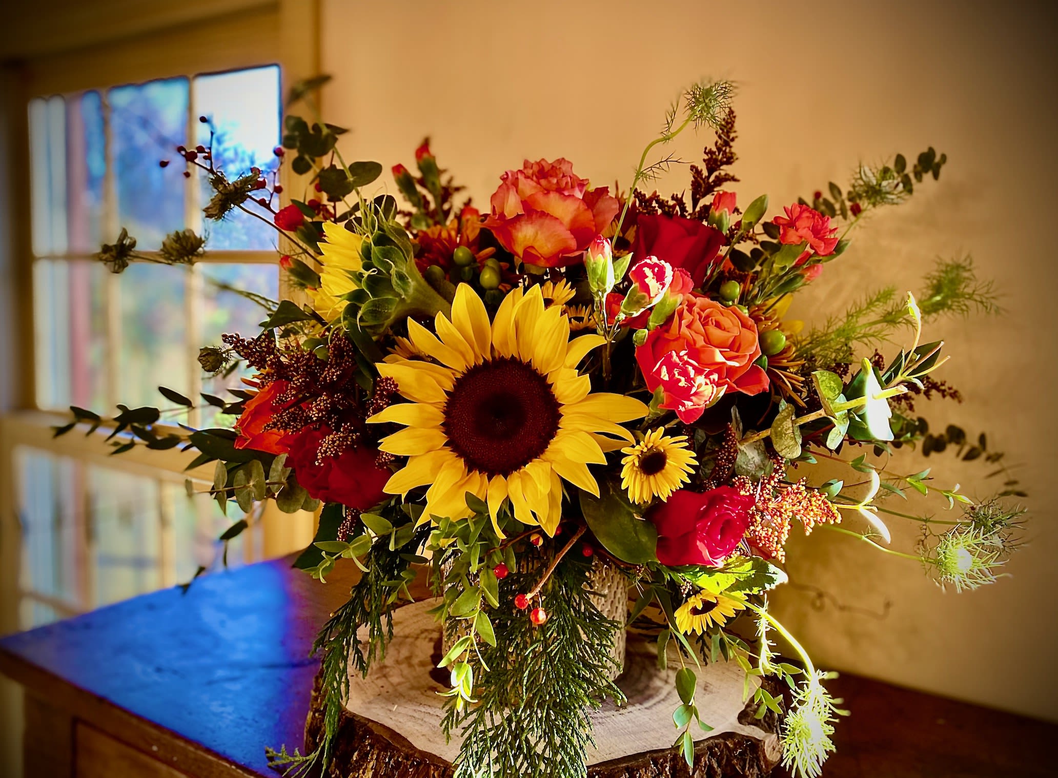 Rustic arrangement of sunflowers, red and orange roses, and daisies in a wood-textured vase