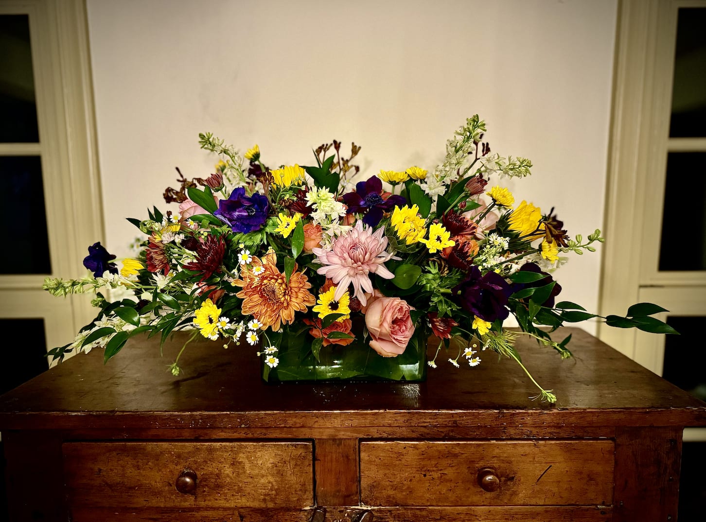 Low rectangular arrangement of mixed yellow, pink, orange, and purple flowers on a wooden dresser