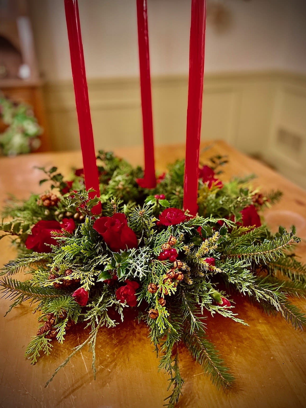 Low evergreen centerpiece with red roses and berries around three tall red taper candles