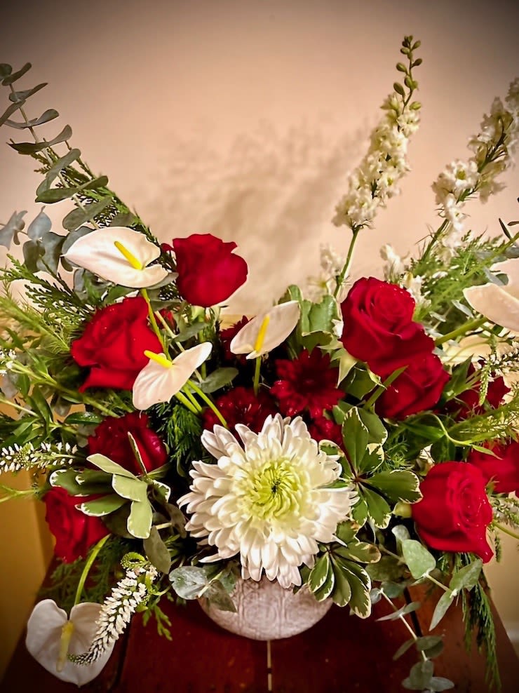 Red roses and white blooms arranged in a round white ceramic vase with assorted foliage.