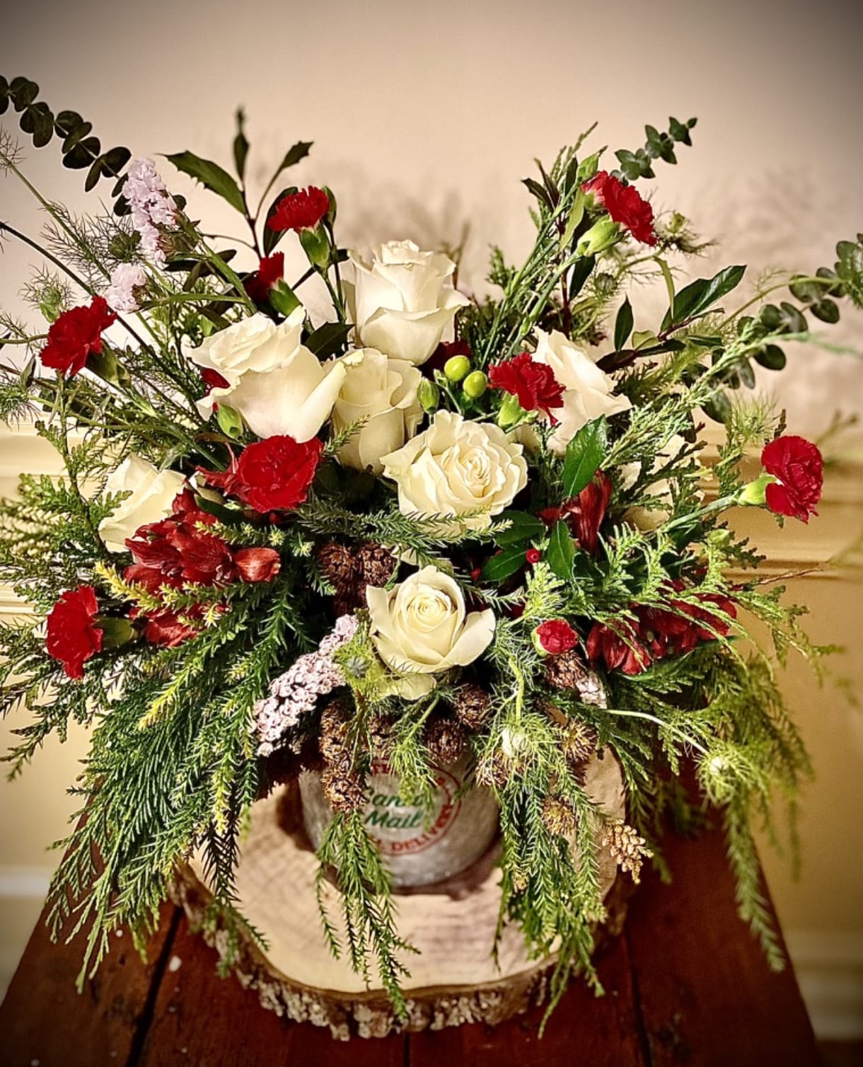 Rustic arrangement of white roses and red carnations with winter greenery in a metal container on a wood slice.