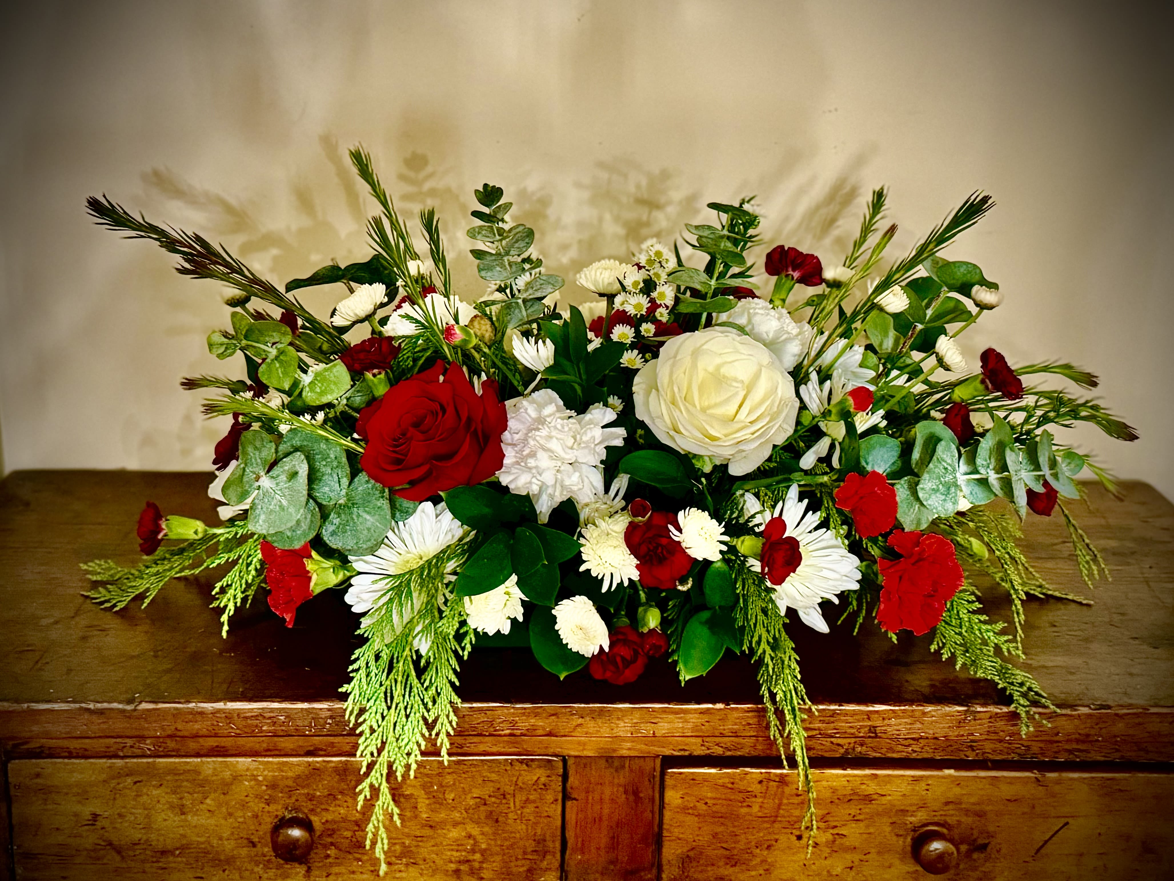 Low red and white flower arrangement with abundant greenery on a wooden dresser