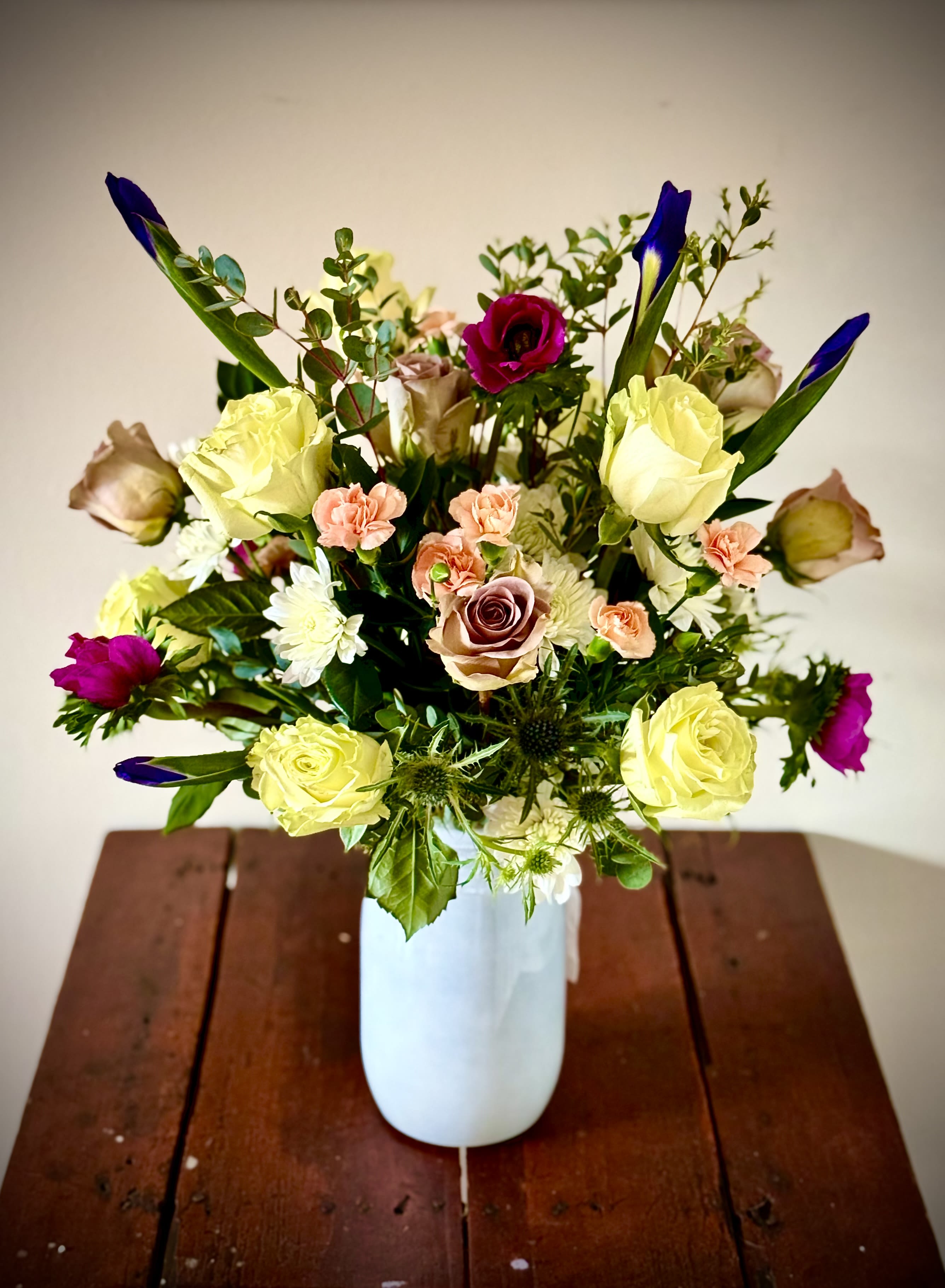 Mixed bouquet of yellow roses, peach carnations, and purple blooms in a white vase on a wooden table