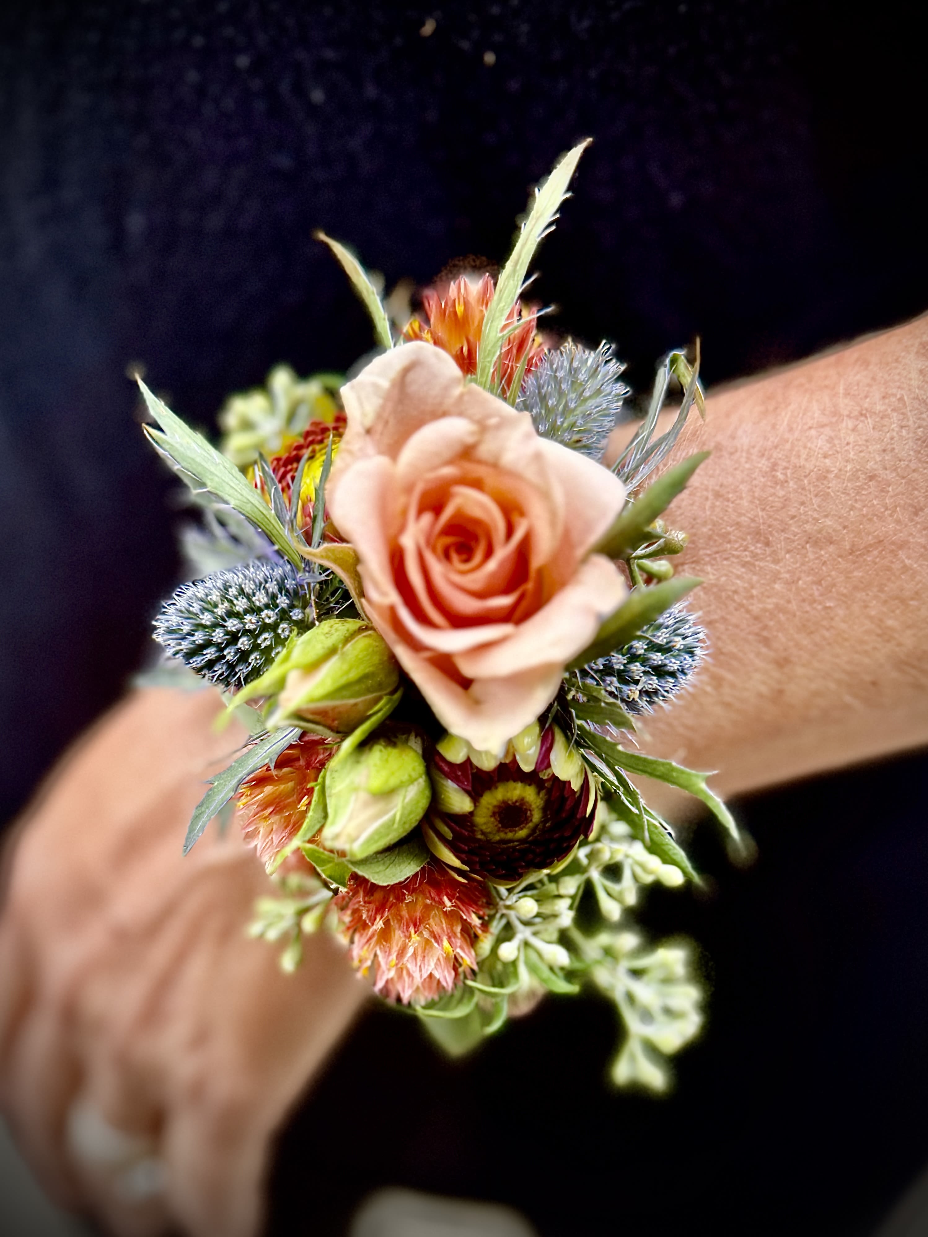 Wrist corsage with a peach rose, spiky blue blooms, and orange accents on a person’s wrist