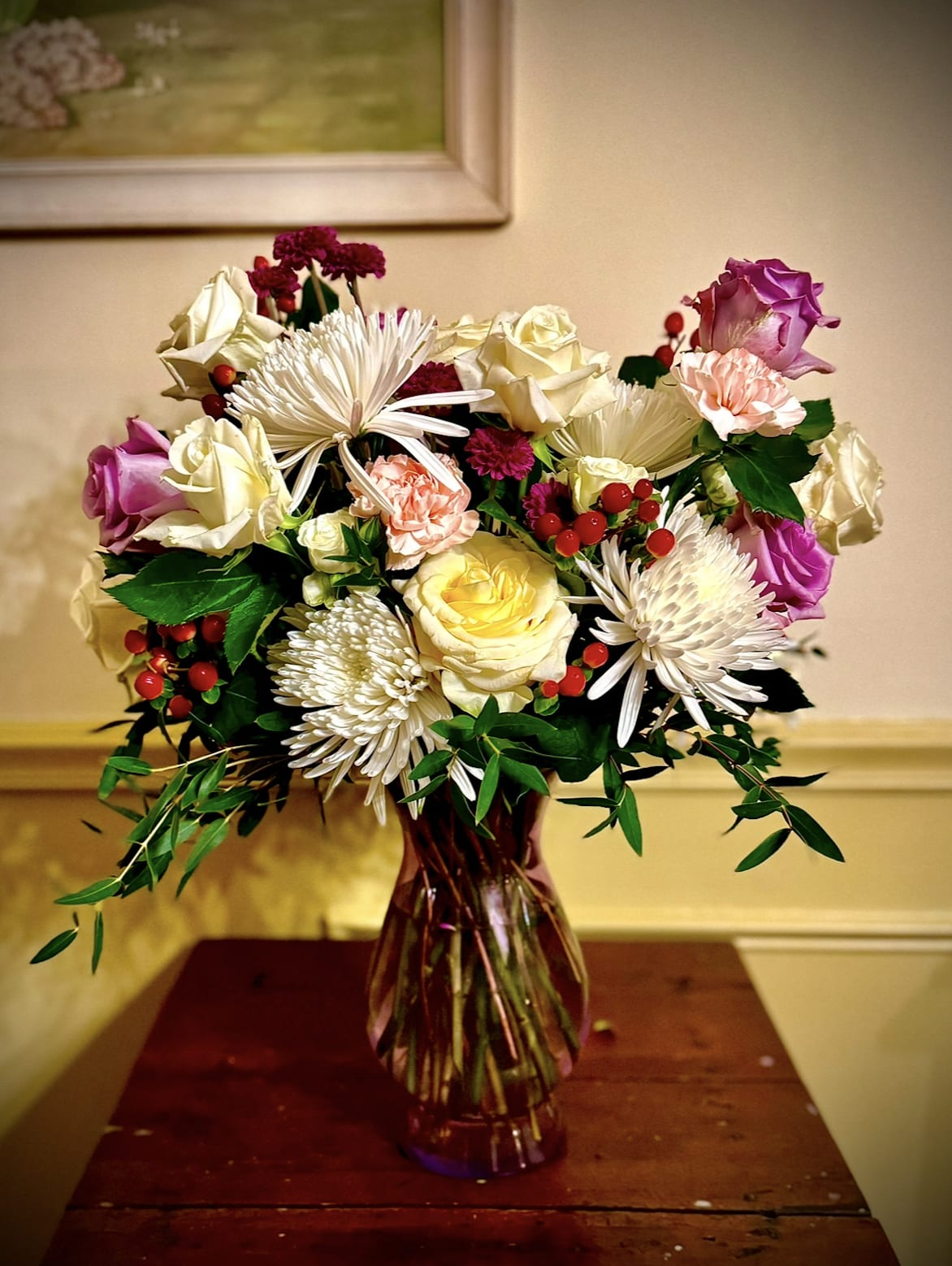 Mixed bouquet of roses, carnations, and white spider mums in a tinted glass vase with red berries