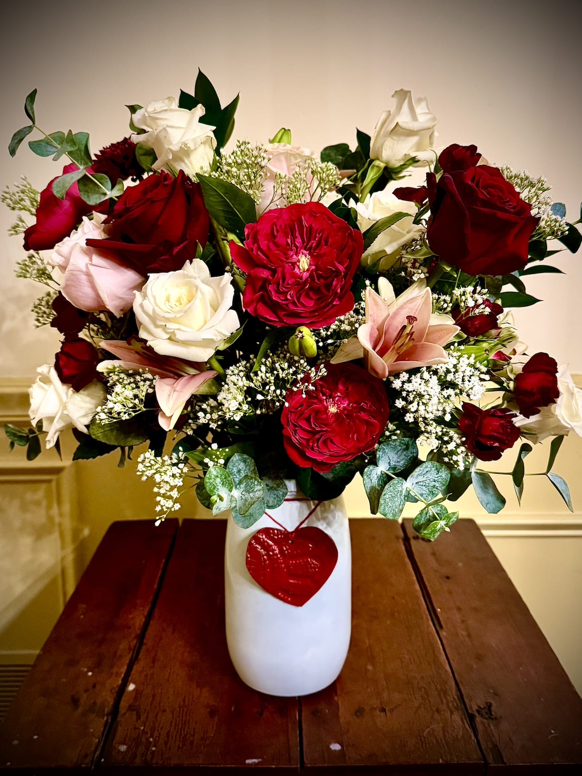 Mixed bouquet of red and white roses with pink lilies in a white vase with a red heart charm.